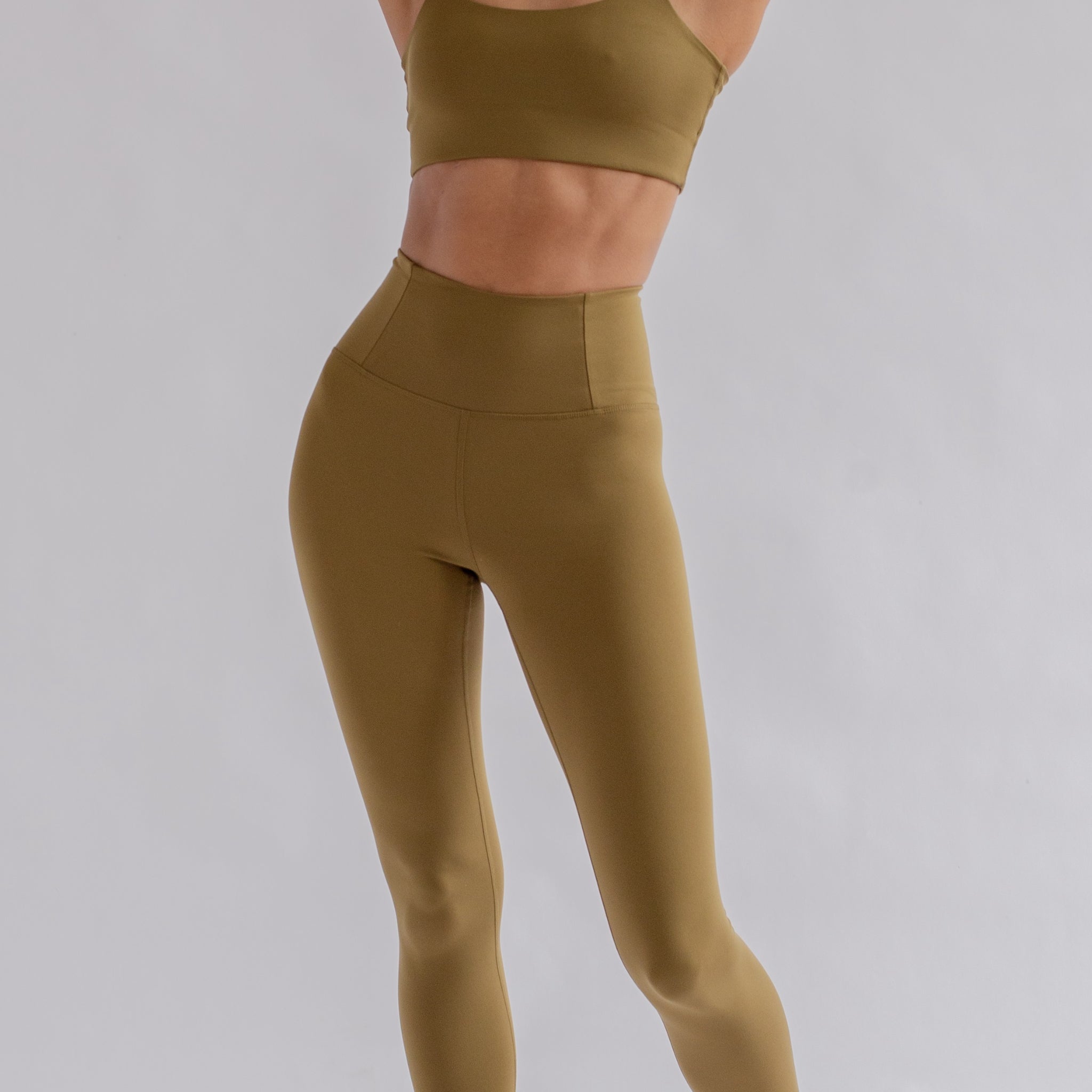 Person in olivegrüner Sportbekleidung, bestehend aus einem Top und einer Leggings, posiert vor grauem Hintergrund.