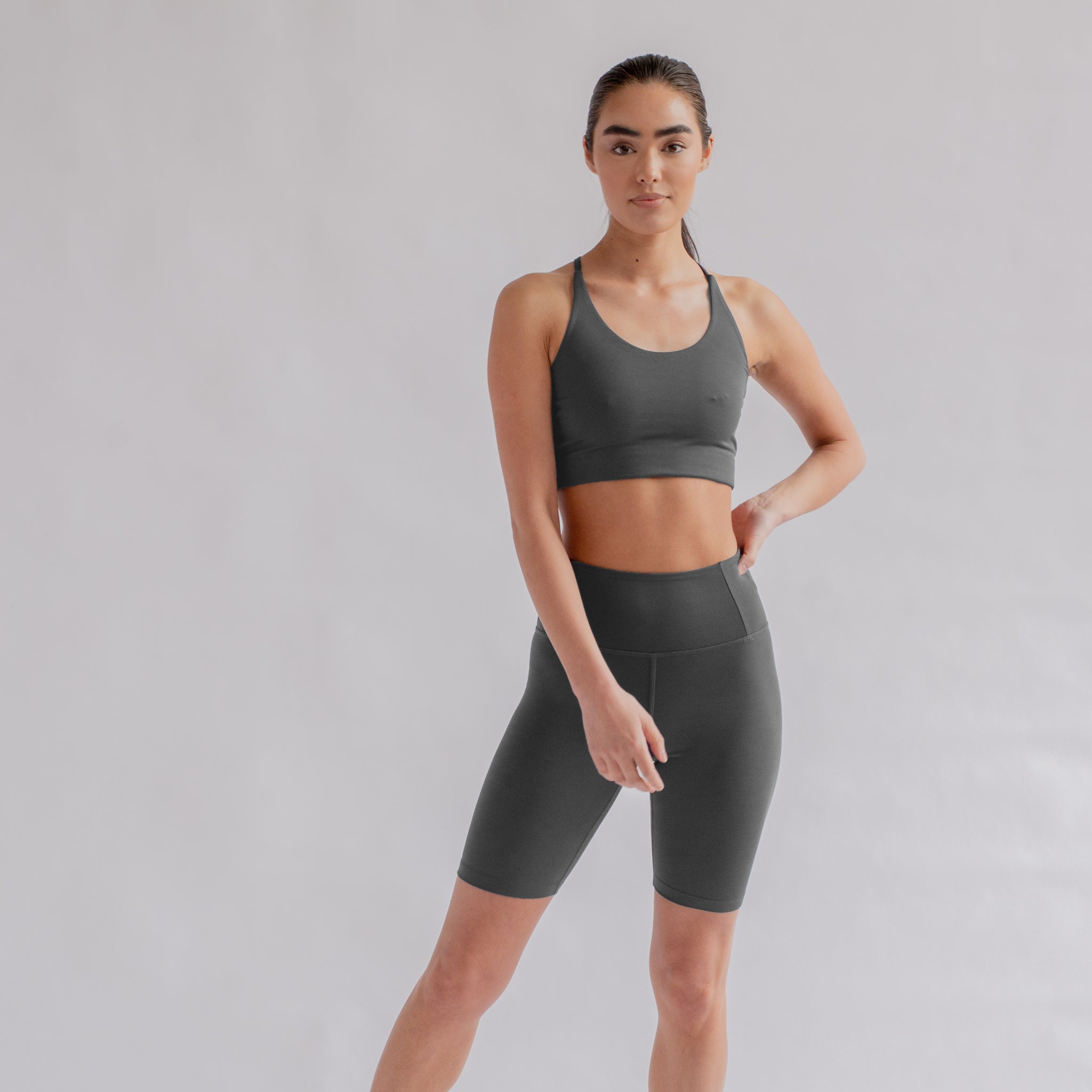 Frau in grauem Sport-BH und Shorts posiert vor neutralem Hintergrund.