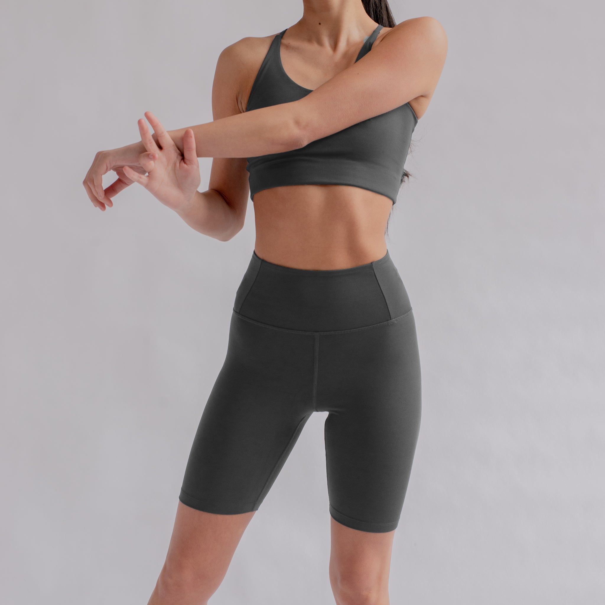Frau in grauem Sport-BH und Shorts streckt ihren Arm vor einem neutralen Hintergrund.