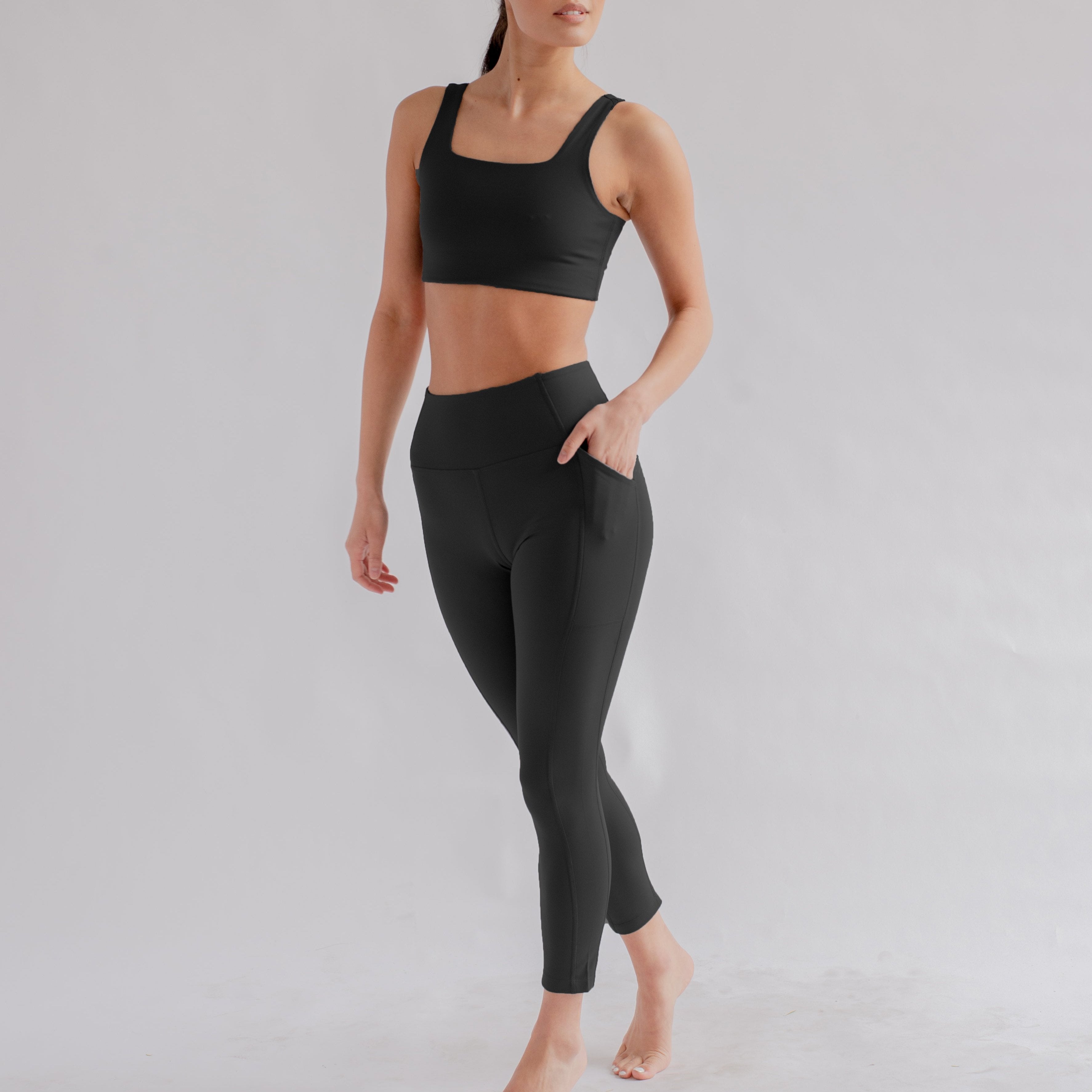 Person in schwarzem Sport-BH und Leggings posiert barfuß vor grauem Hintergrund.