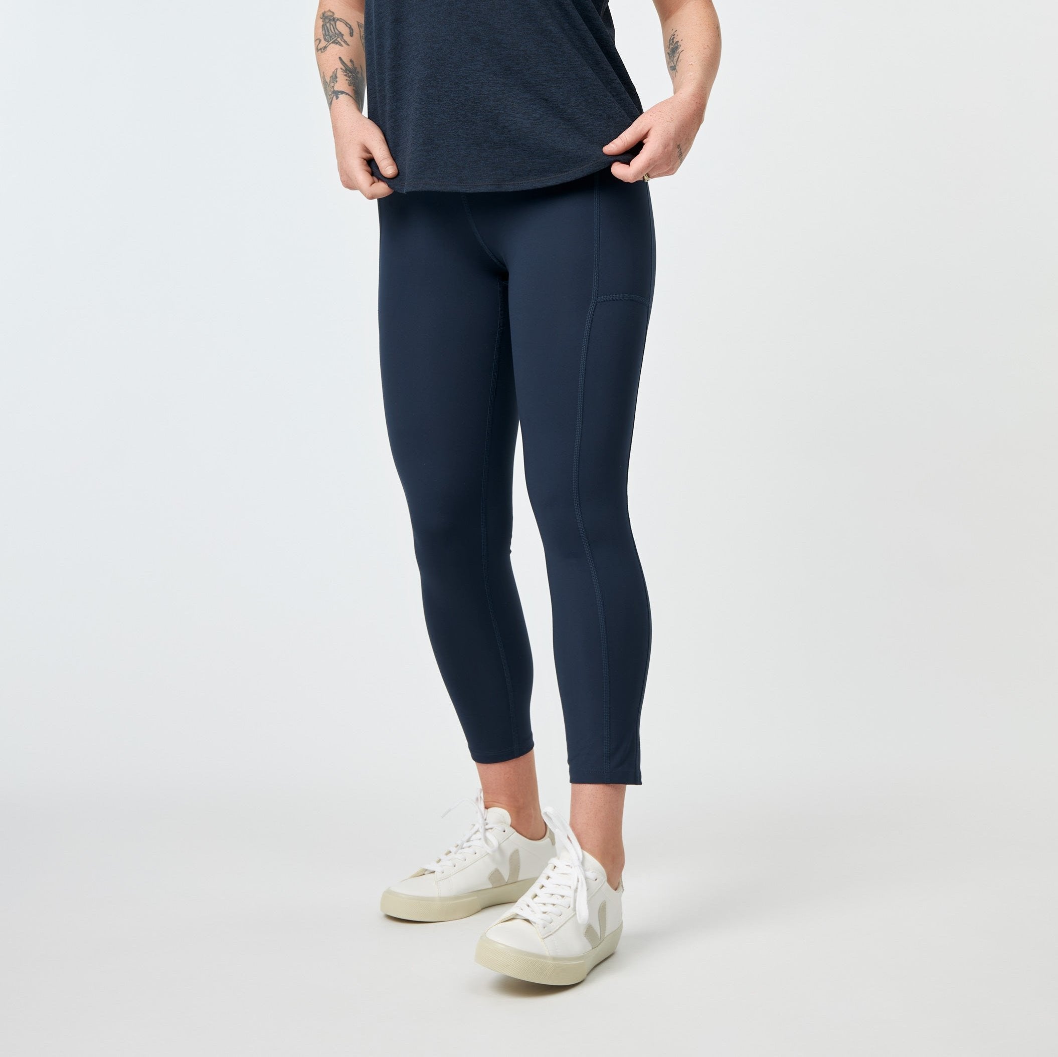 Person in blauer Leggings und weißem T-Shirt, steht mit weißen Sneakers auf grauem Hintergrund.