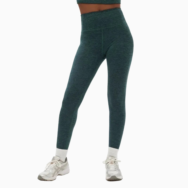 Eine Person, die blaugrüne Leggings, weiße Socken und silberne Sportschuhe trägt, steht vor einem schlichten weißen Hintergrund. Ihr Oberkörper befindet sich größtenteils außerhalb des Bildausschnitts.