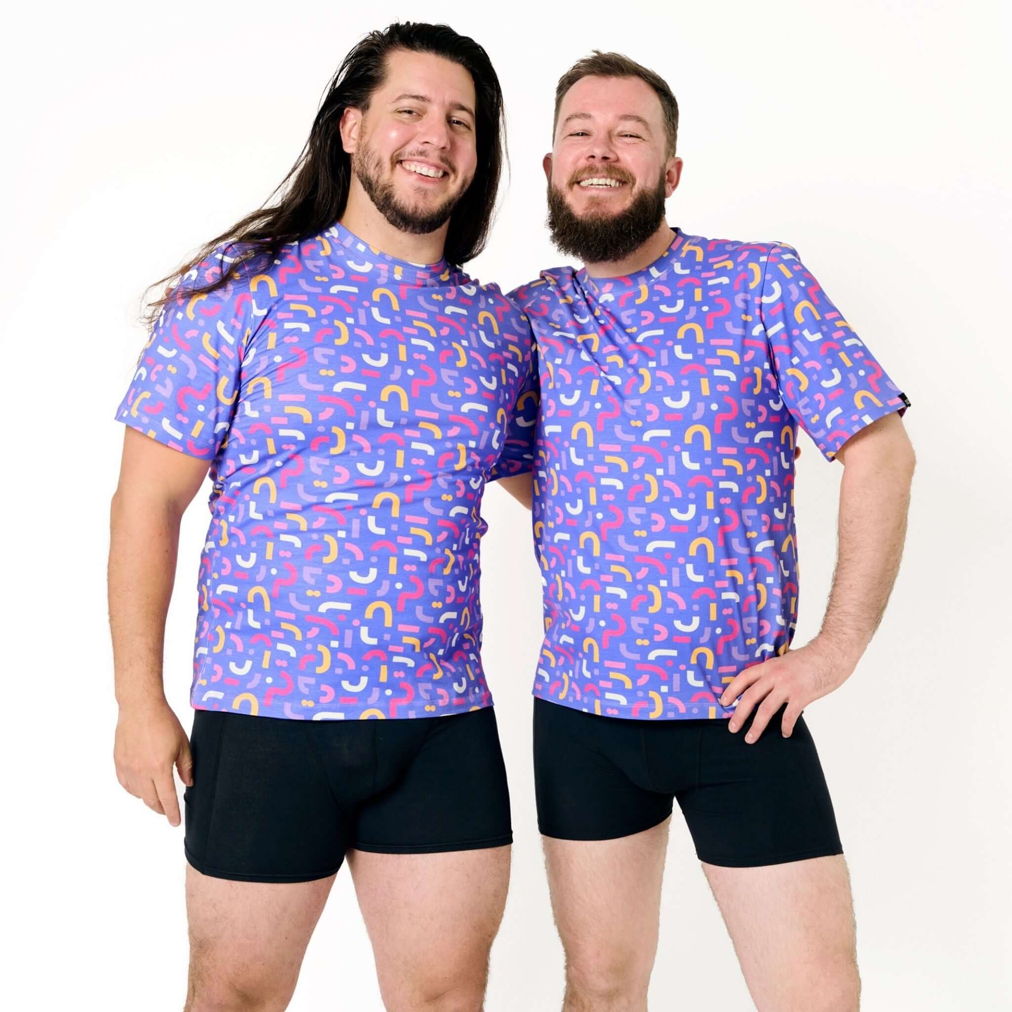 Zwei Männer in bunten T-Shirts und schwarzen Shorts lächeln und posieren vor einem weißen Hintergrund.