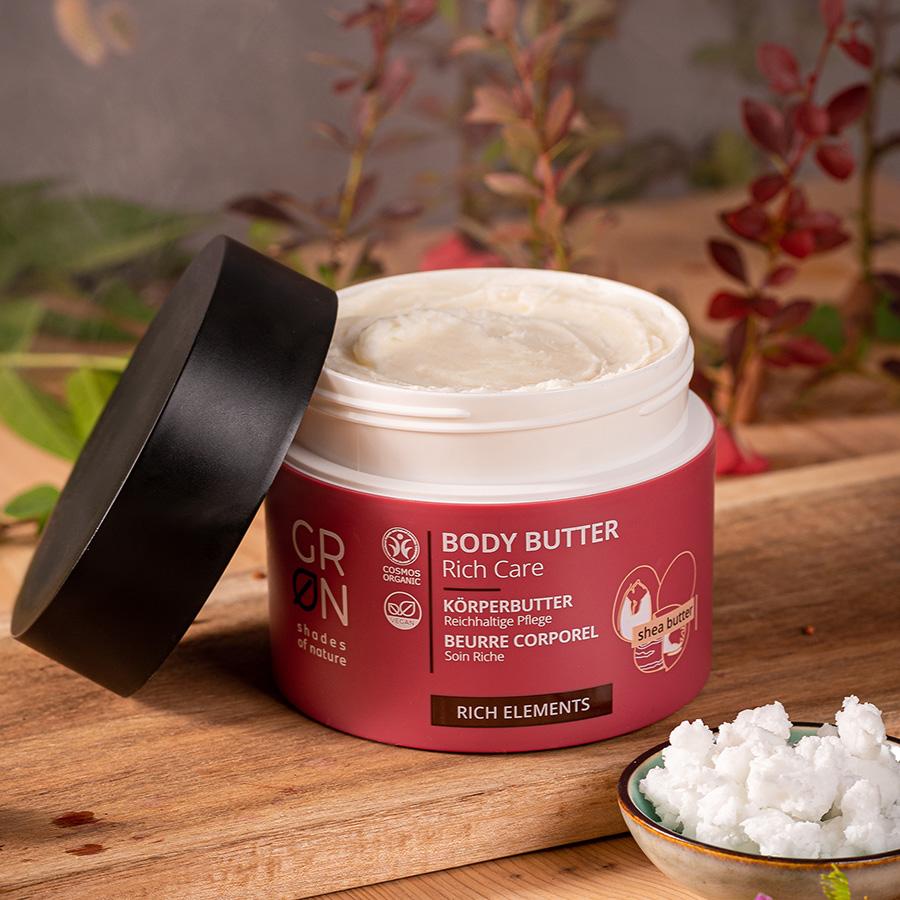 Ein Tiegel GRN Body Butter Rich Care steht ohne Deckel auf einer Holzfläche, umgeben von Grünzeug und einer kleinen Schale mit Sheabutter. Das Etikett hebt hervor, dass sie reich an Sheabutter ist und sich für die Körperpflege eignet.