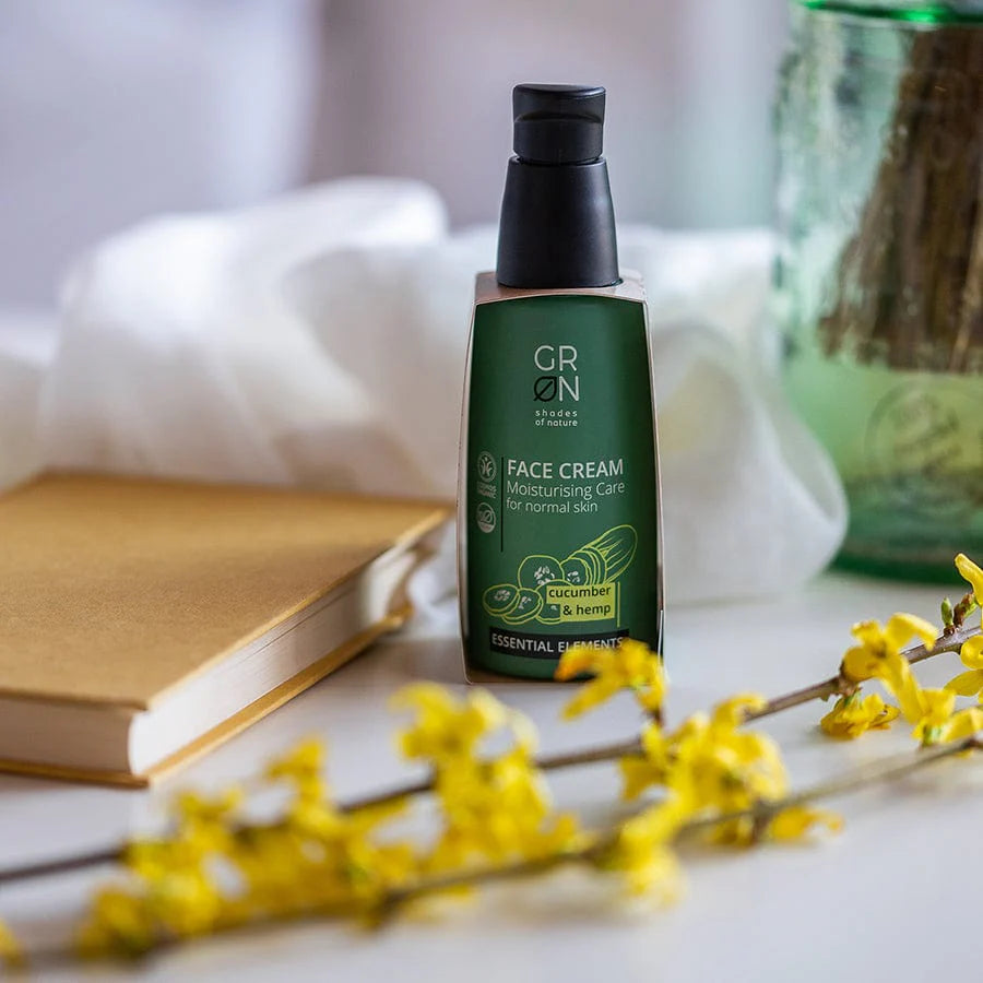 Eine grüne Flasche GRN-Gesichtscreme steht auf einem Tisch neben einem geschlossenen Buch, mit einem verschwommenen Glastiegel und gelben Blumen im Vordergrund. Auf dem Etikett der Flasche steht: "Gurke und Hanf für normale Haut".