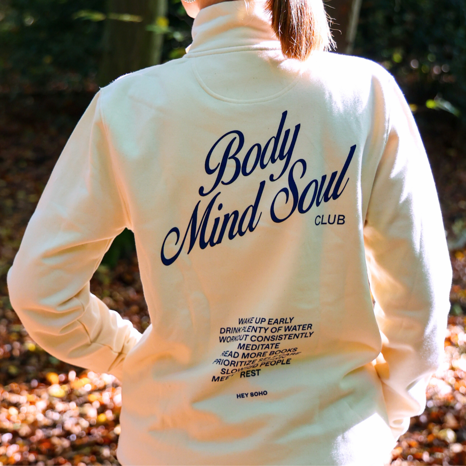 Person in white Pullover mit Aufschrift "Body Mind Soul Club" stehend im Wald, Sonnenlicht scheint durch die Bäume.