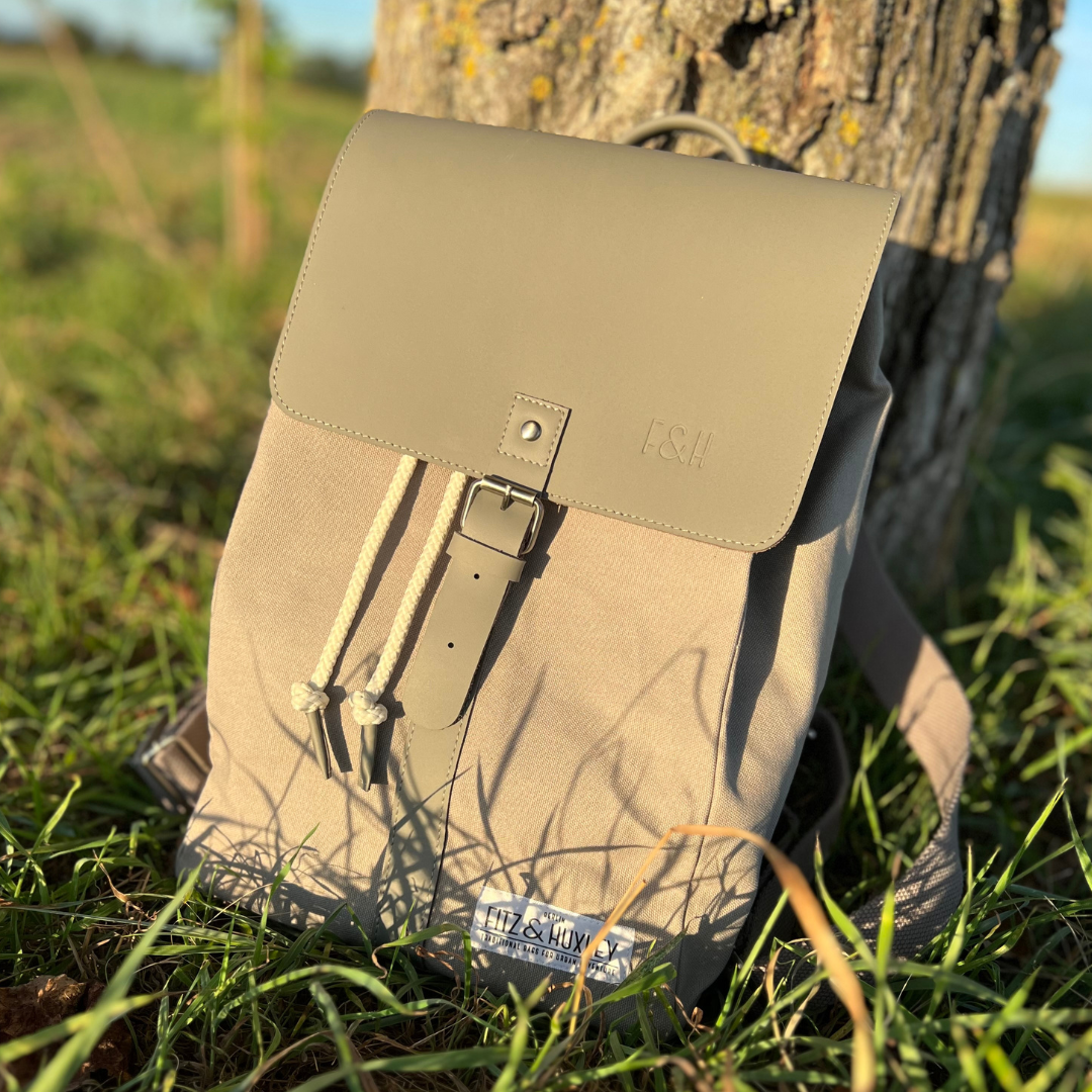 Beiger Rucksack mit Kordelzug und Lederriemen lehnt an einem Baumstamm im Gras bei Sonnenlicht.