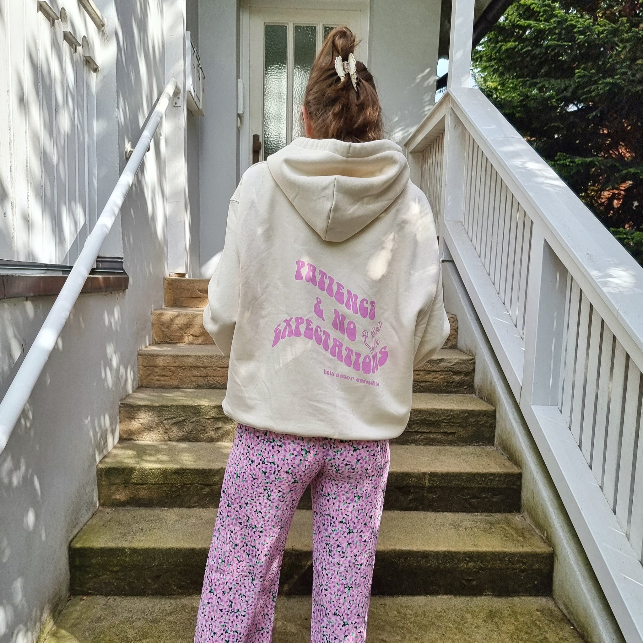 Person in cremefarbenem Hoodie mit rosa Aufschrift und bunter Hose steht auf einer Treppe vor einer Tür.