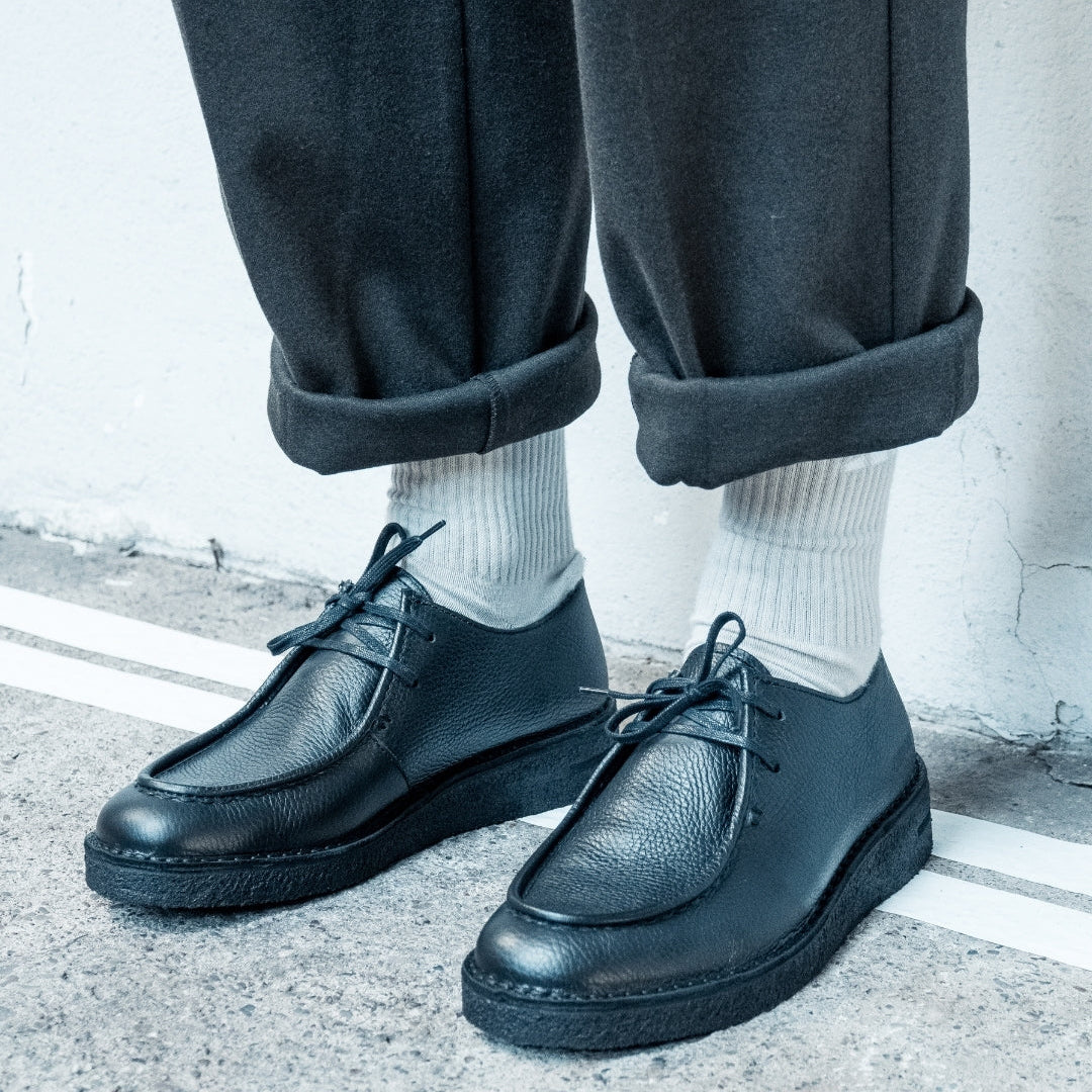 Eine Person mit schwarzen Lederschnürschuhen, weißen Socken und einer dunklen, hochgekrempelten Hose steht auf einer Betonfläche neben einer weißen Linie.