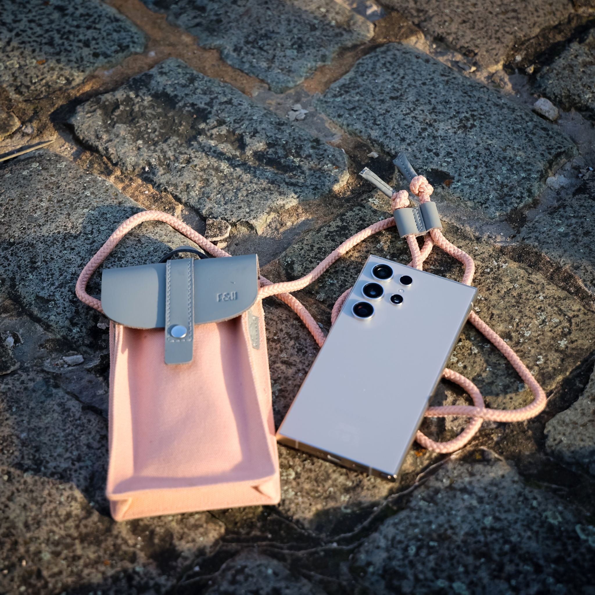 Smartphone mit vier Kameralinsen neben einer rosa Stofftasche mit grauem Verschluss auf Kopfsteinpflaster.