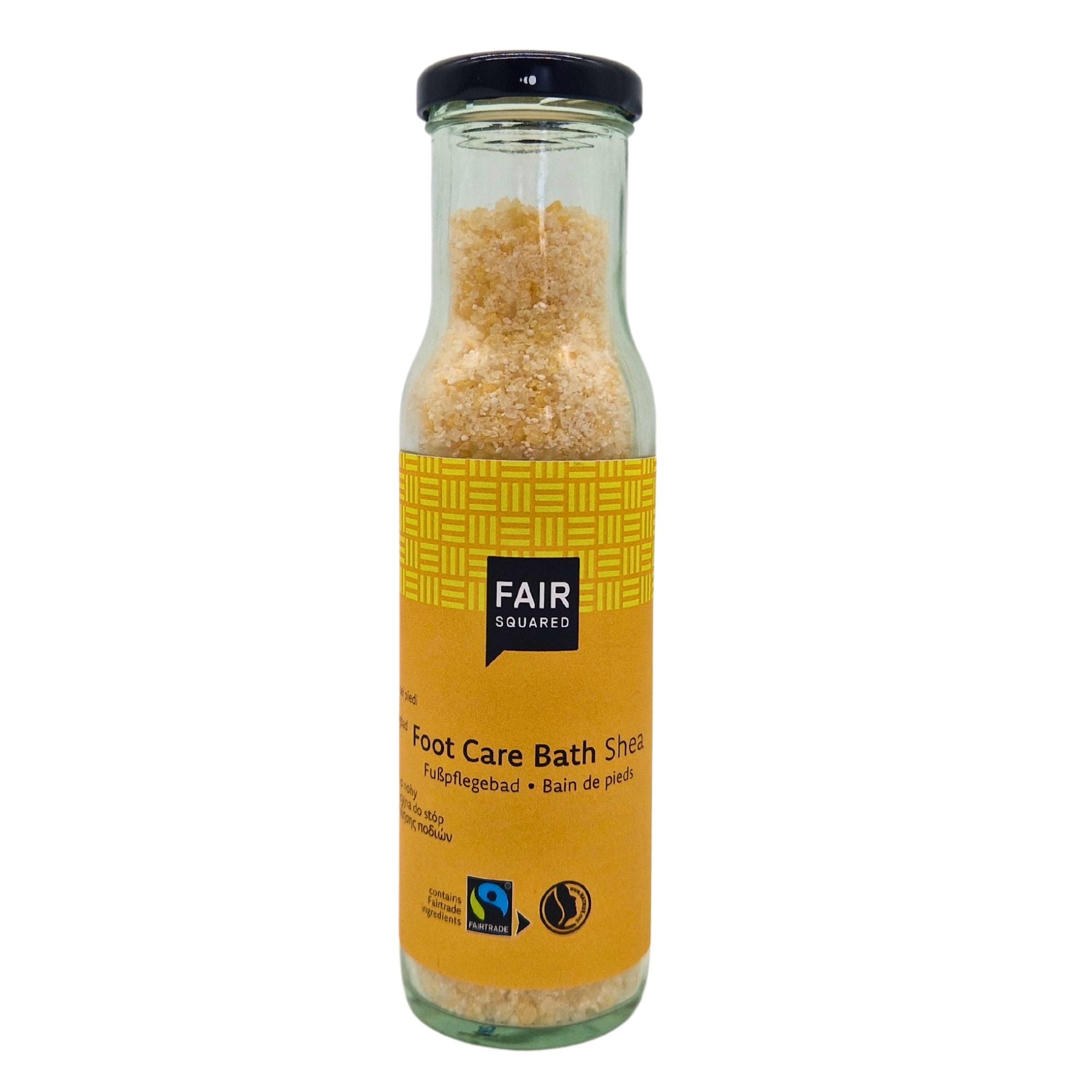 Eine Glasflasche gefüllt mit gelbem Badesalz mit der Aufschrift Fair Squared Foot Care Bath Shea mit Fairtrade- und Vegan-Logos auf dem gelben Etikett. Die Flasche hat einen schwarzen Twist-off-Verschluss.