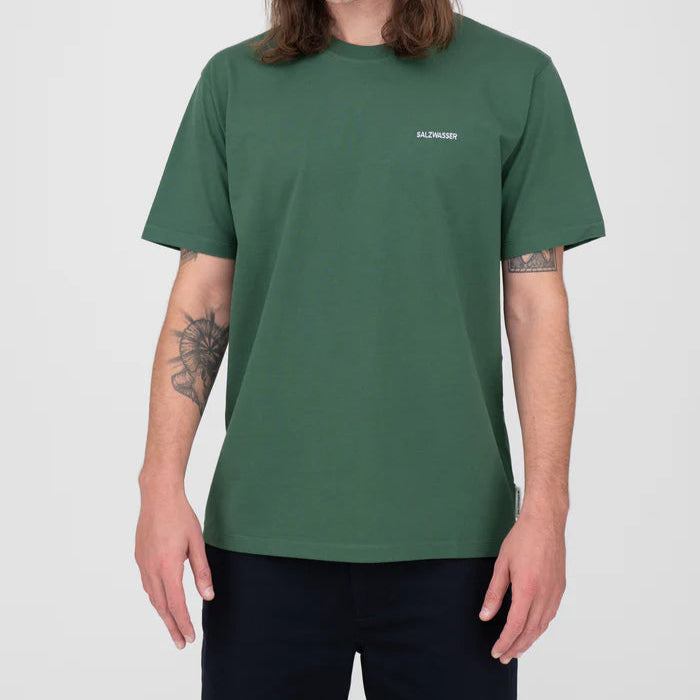 Eine Person mit langen Haaren trägt ein dunkelgrünes, kurzärmeliges T-Shirt mit der Aufschrift SALZWASSER" in kleiner weißer Schrift auf der linken Brust, dazu eine dunkle Hose. Die Arme sind seitlich entspannt und zeigen eine Tätowierung auf einem Unterarm.