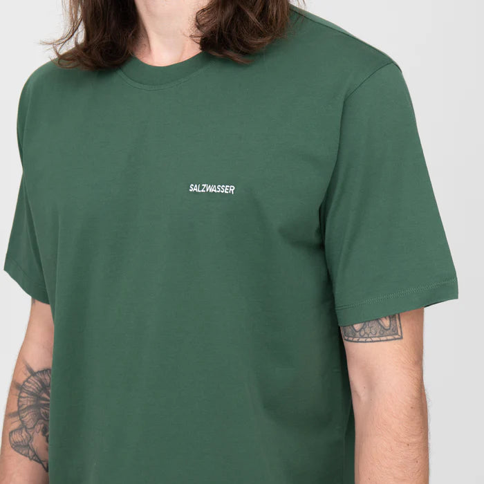 Eine Person mit langen braunen Haaren trägt ein grünes T-Shirt mit dem Aufdruck SALZWASSER in kleinen weißen Buchstaben auf der Brust. Der linke Arm der Person hat sichtbare Tätowierungen. Der Hintergrund ist einfarbig und hell gefärbt.