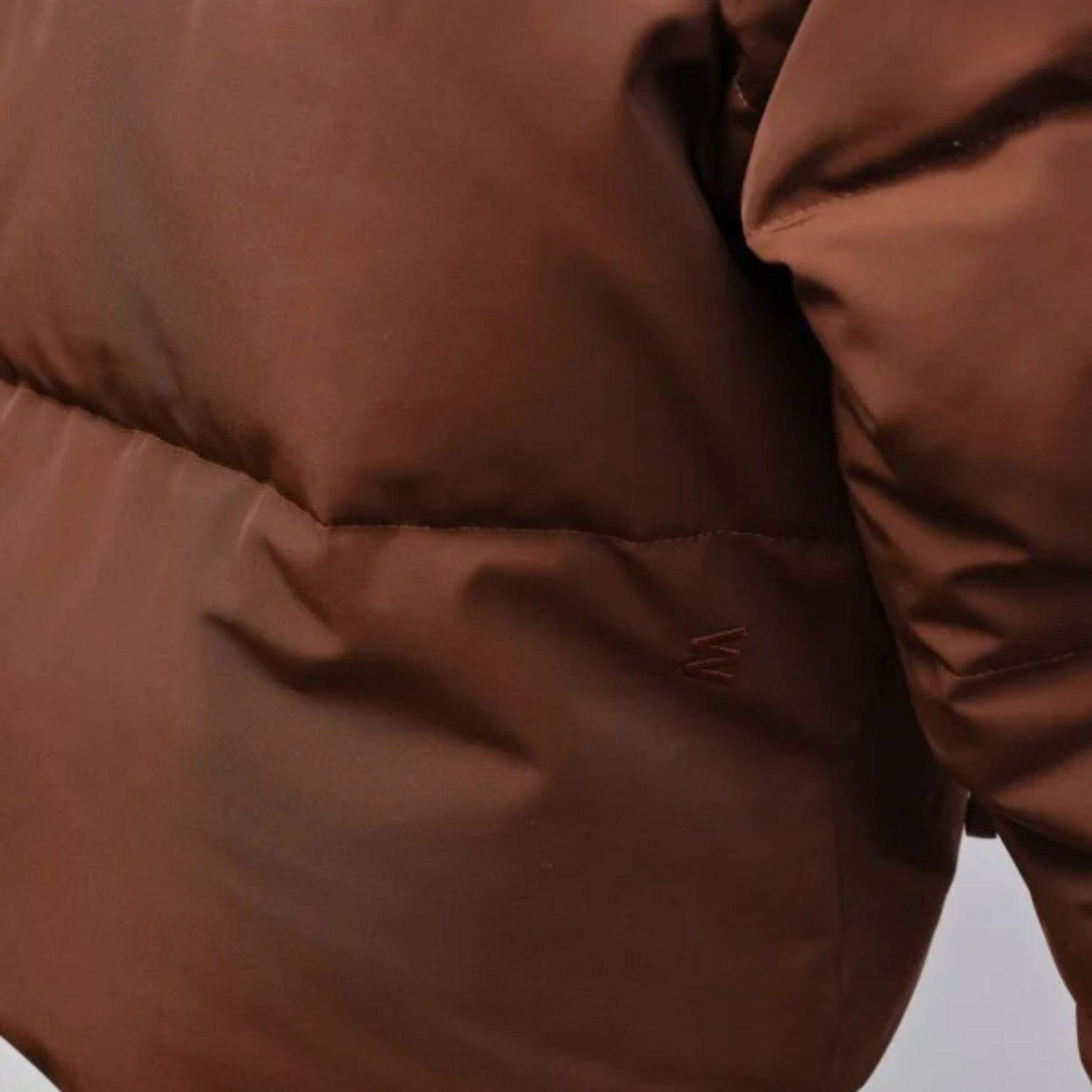 Nahaufnahme einer braunen Steppjacke mit sichtbarer Naht und dezentem Logo auf der linken Seite.