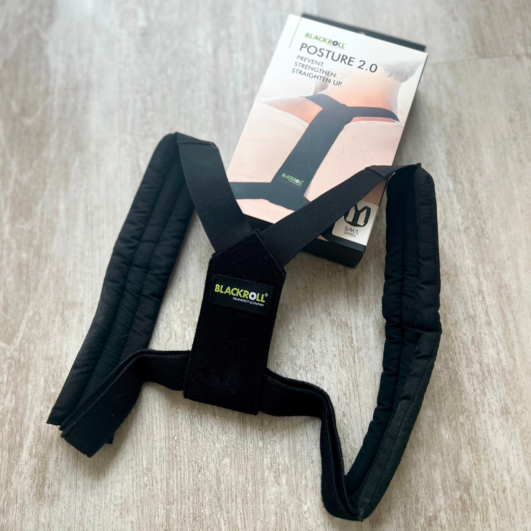 Schwarze Haltungskorrekturhilfe "Blackroll Posture 2.0" neben ihrer Verpackung auf einem Holzboden.