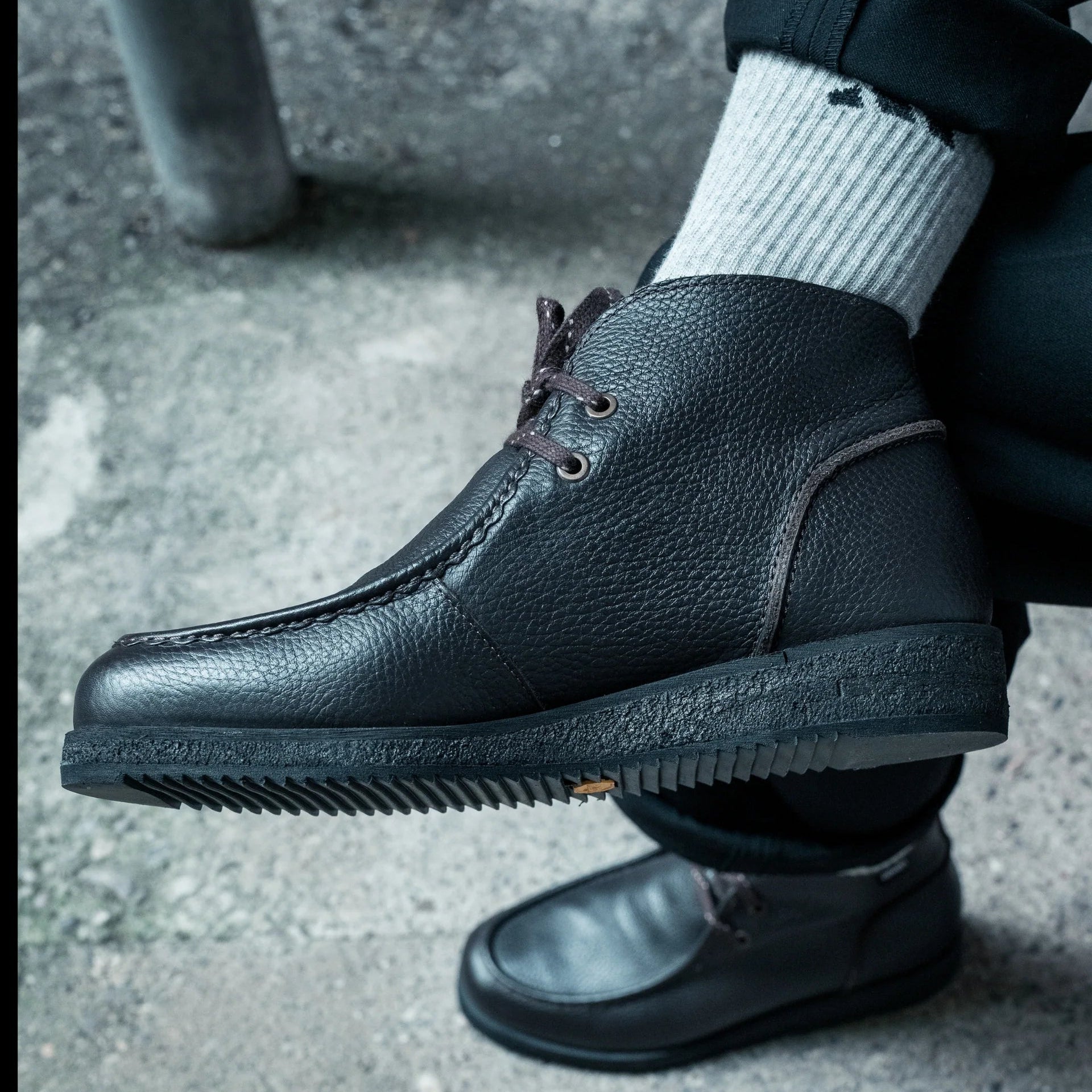Eine Person, die hellgraue Socken und schwarze strukturierte Lederschuhe trägt, sitzt mit einem übereinander geschlagenen Bein auf einer Betonfläche.
