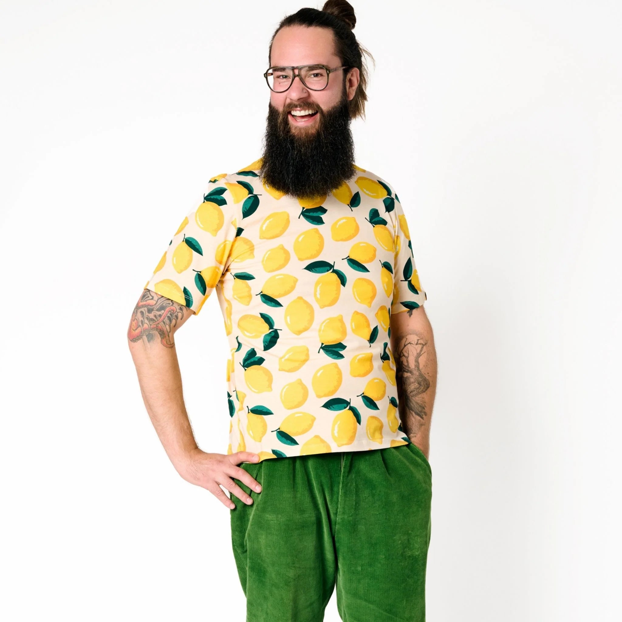 Mann mit Brille und Bart trägt ein gelbes T-Shirt mit Zitronenmuster und grüne Hose, lächelnd vor weißem Hintergrund.