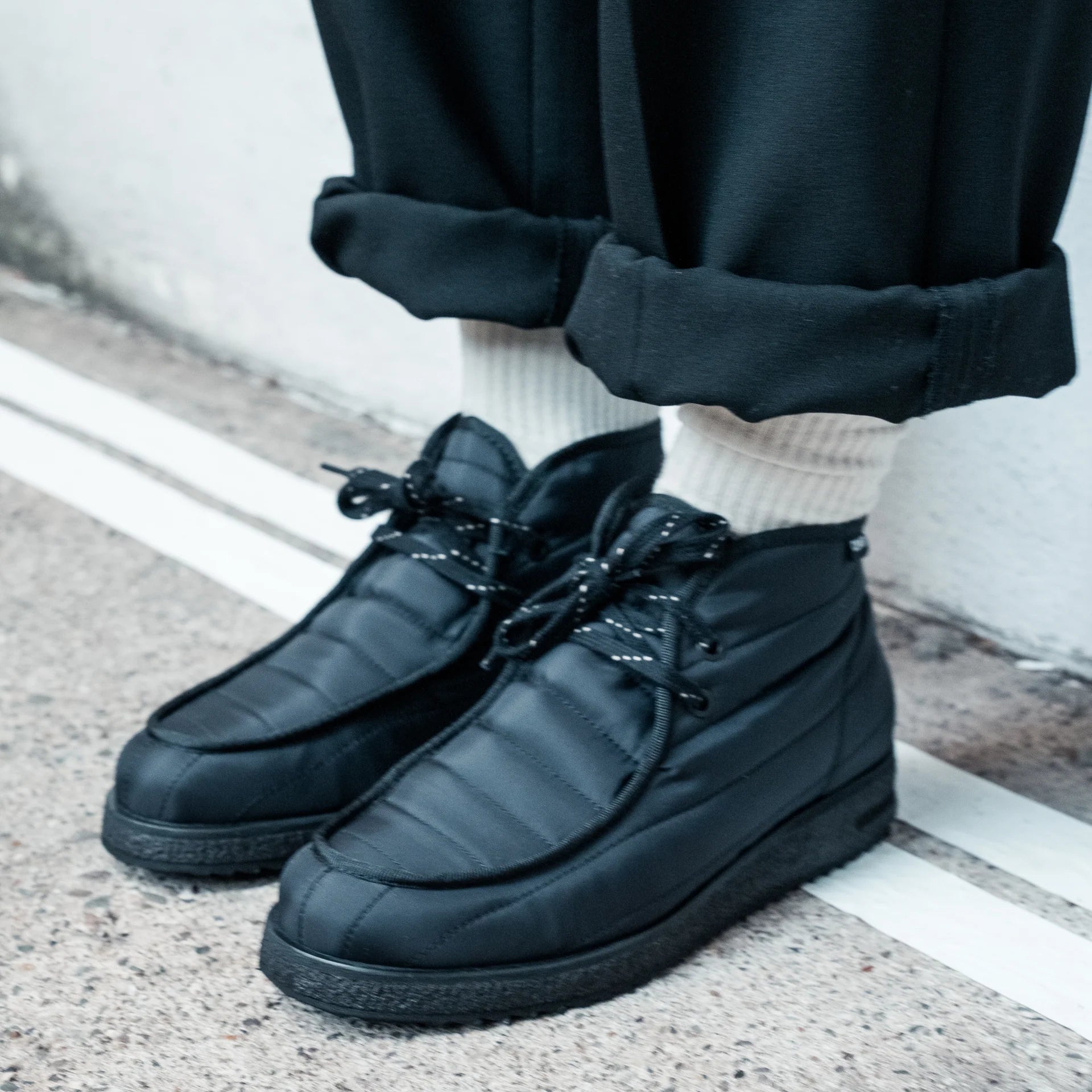 Eine Person, die schwarze, isolierte Schnürschuhe mit weißen Socken und eine schwarze, mit Manschetten versehene Hose trägt, steht auf einer Betonfläche in der Nähe einer weißen Linie und Wand.