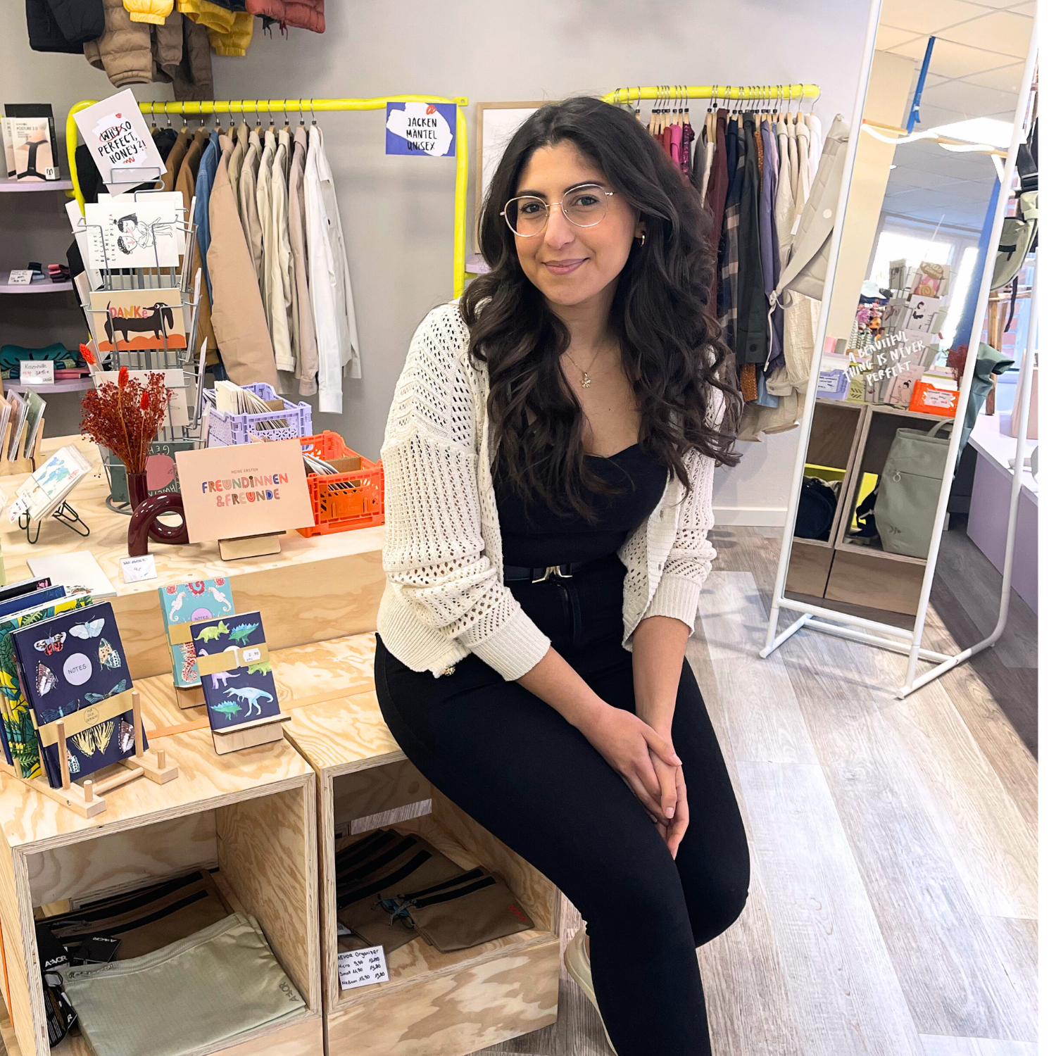 Frau mit Brille sitzt in einem kleinen Laden vor Regalen mit Karten und Kleidung.