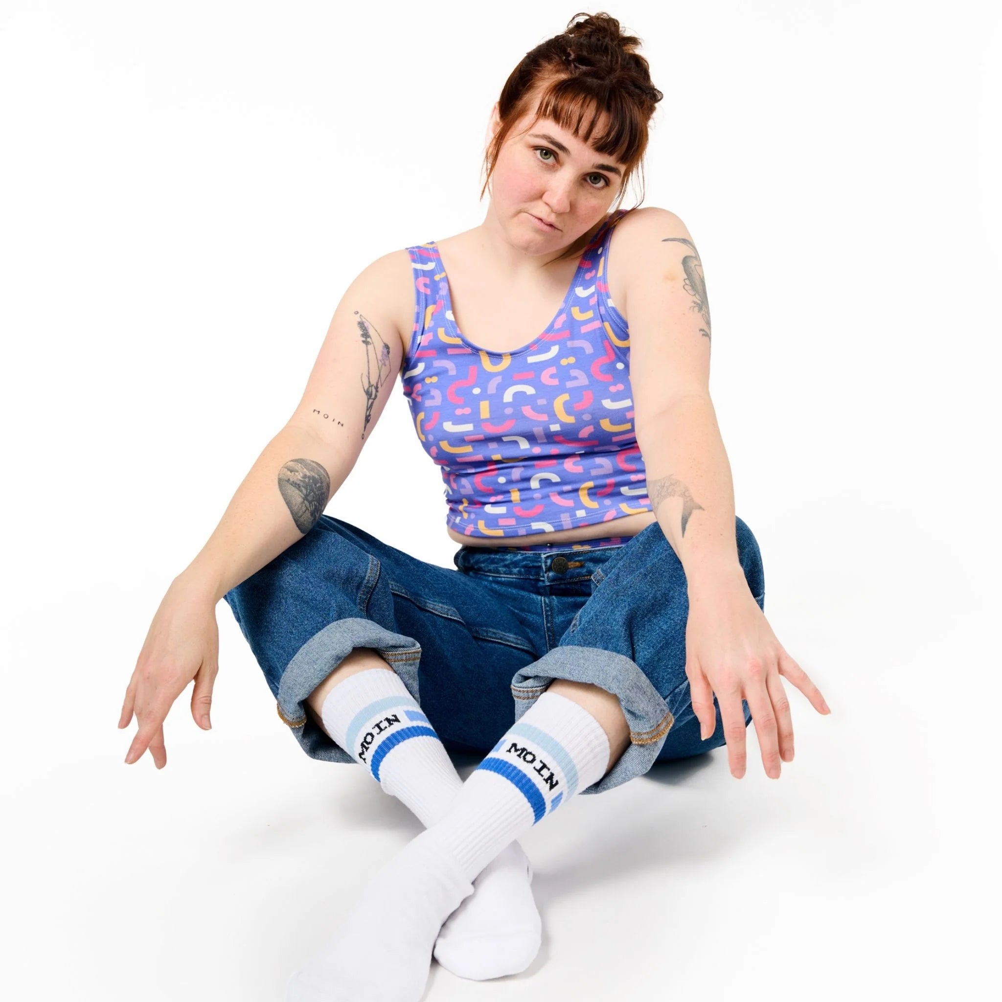 Frau in gemustertem Top und gekrempelter Jeans sitzt auf weißem Hintergrund, Arme entspannt auf den Knien ruhend.