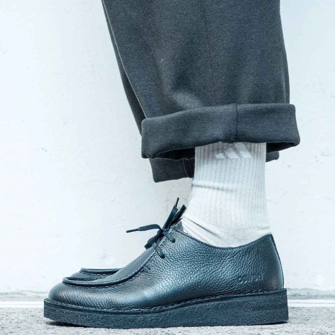 Eine Person, die eine dunkle, hochgekrempelte Hose, weiße Socken und schwarze, strukturierte Lederschnürschuhe trägt, steht auf einer Betonfläche vor einem hellen Hintergrund.