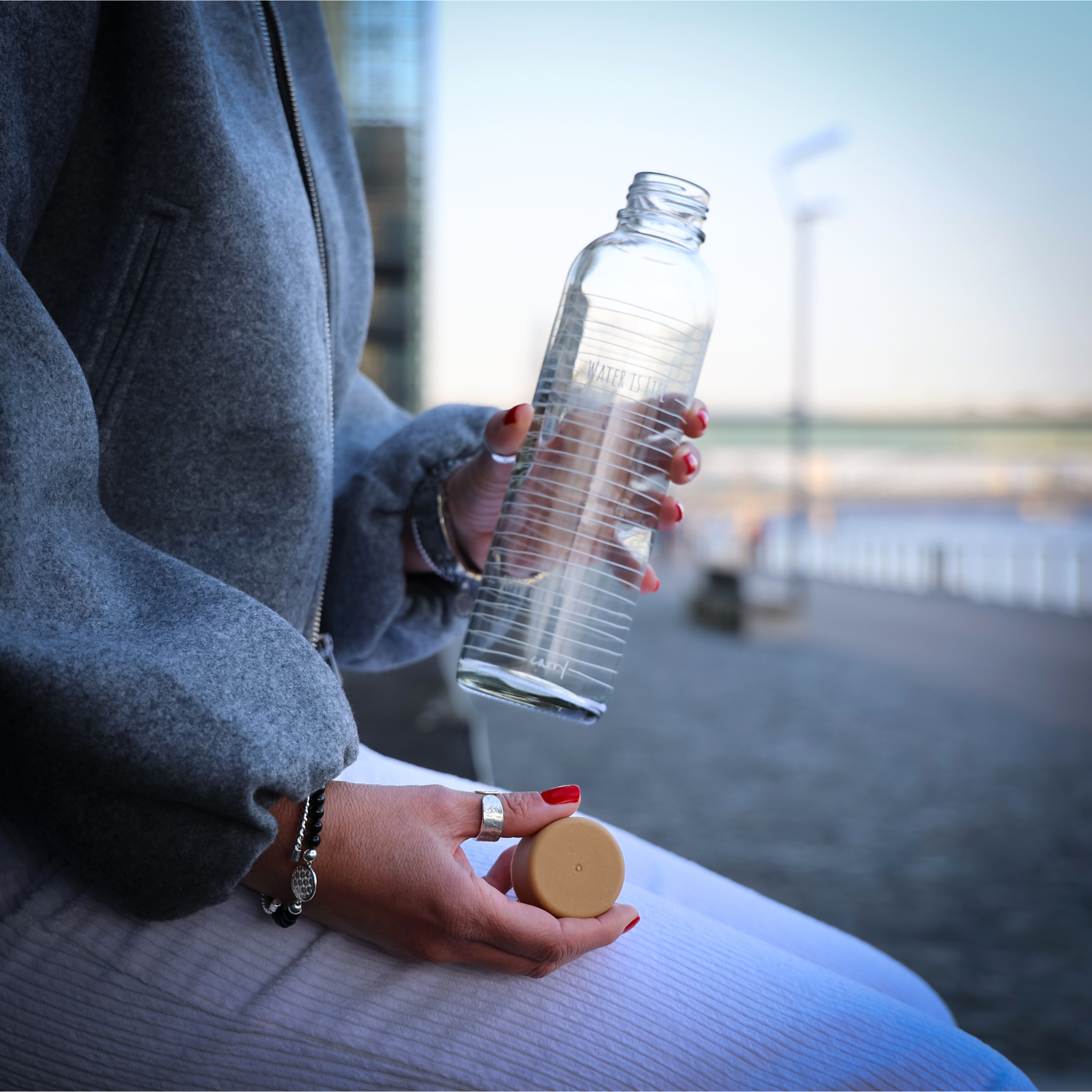 Eine Person in grauer Jacke hält eine leere Glasflasche mit einem beigen Deckel in der Hand.