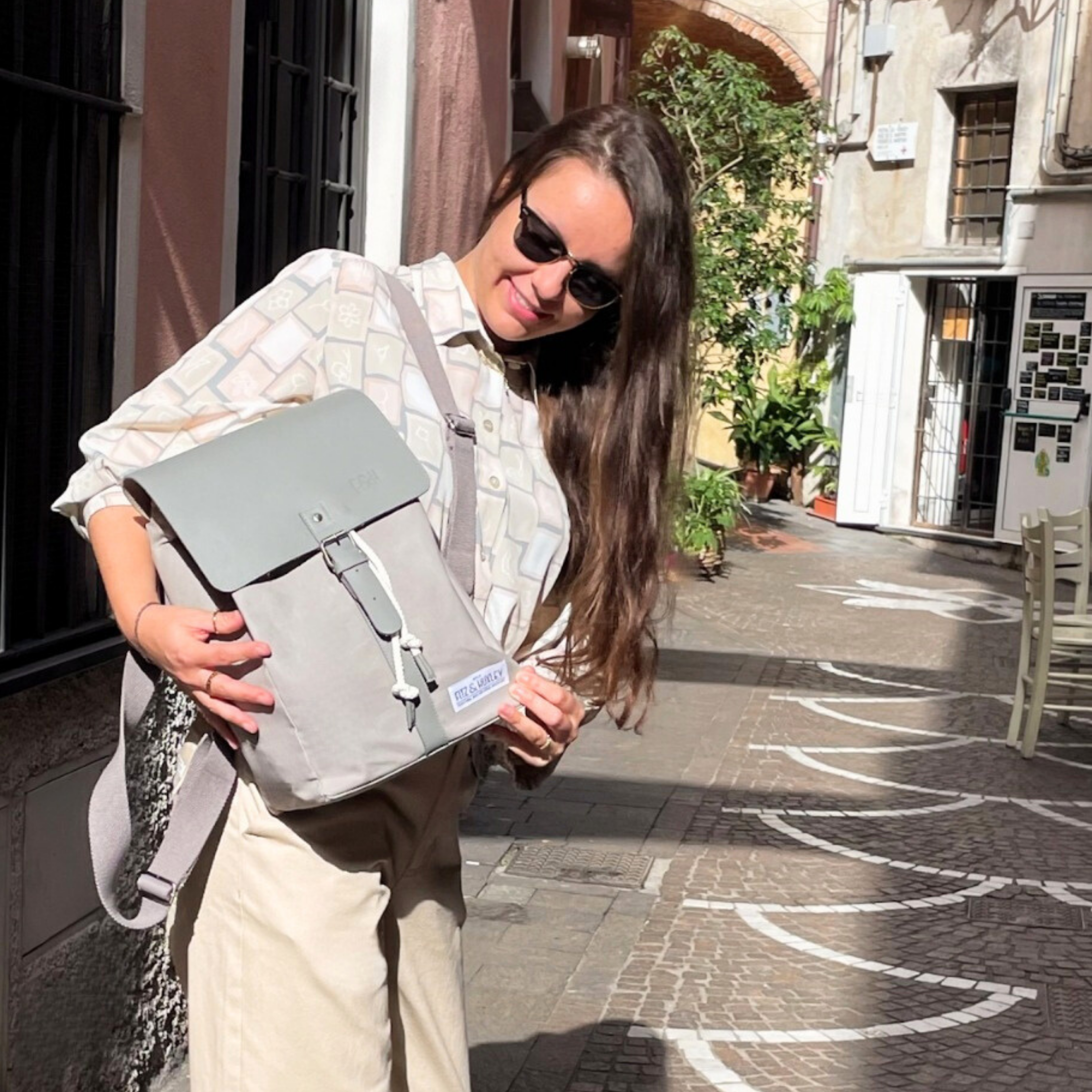 Frau mit Sonnenbrille hält einen hellgrauen Rucksack in einer sonnigen Gasse.