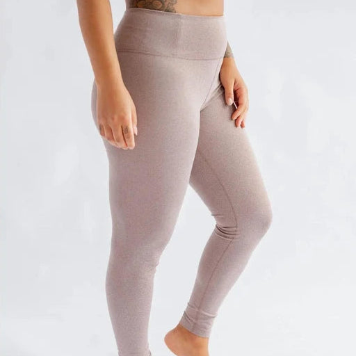 Eine Person, die barfuß auf einer weißen Fläche steht und einen passenden, hellvioletten Sport-BH und Leggings trägt. Die Arme liegen entspannt an der Seite, und auf der linken Hüfte und dem Oberschenkel ist eine Tätowierung zu sehen.