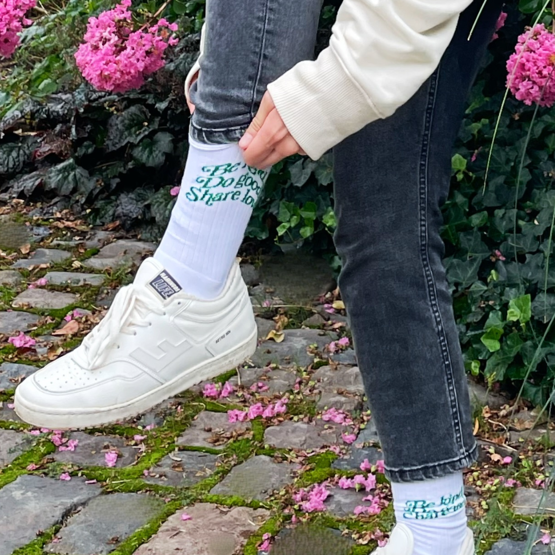 Person trägt weiße Sneaker und Socken mit dem Text "Be Kind, Do Good, Share Love" in einem Garten mit Blumen.