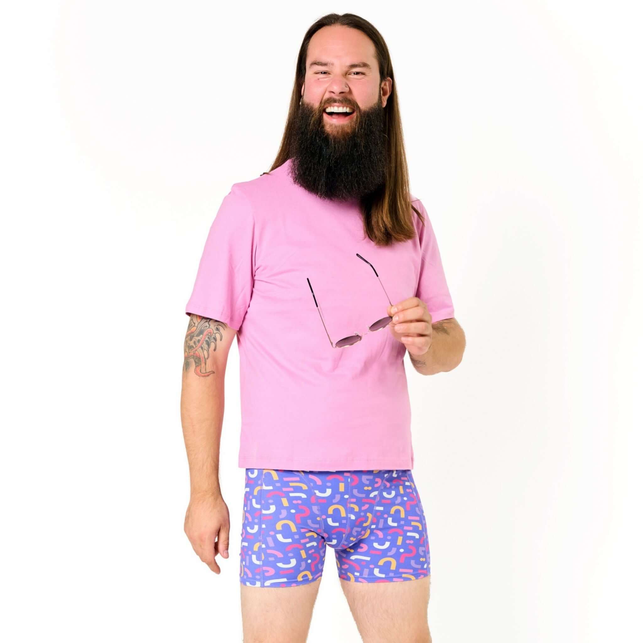 Mann mit langem Haar und Vollbart trägt ein rosa T-Shirt und bunte Boxershorts, hält eine Brille in der Hand.
