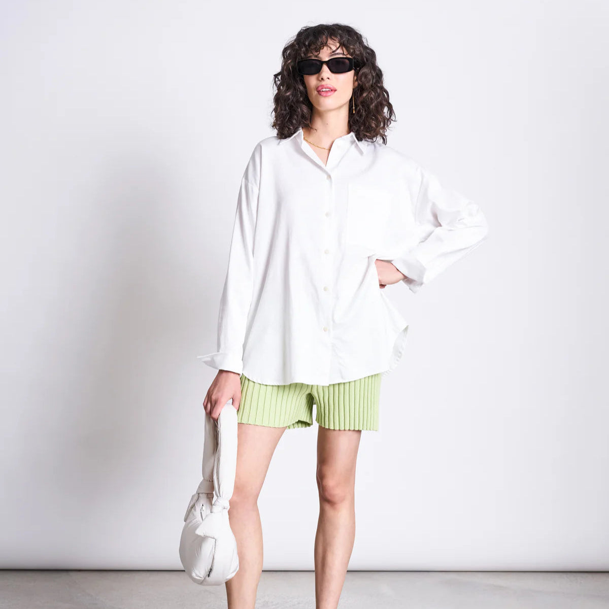 Frau in weißem Hemd und hellgrünen Shorts posiert mit Sonnenbrille und weißer Handtasche vor weißem Hintergrund.