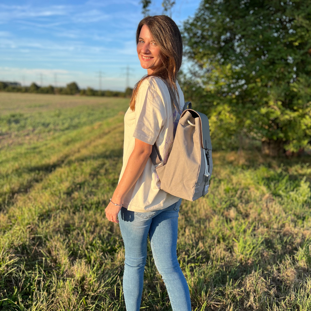 Eine Frau steht mit einem Rucksack auf einer Wiese, im Hintergrund Bäume und blauer Himmel.