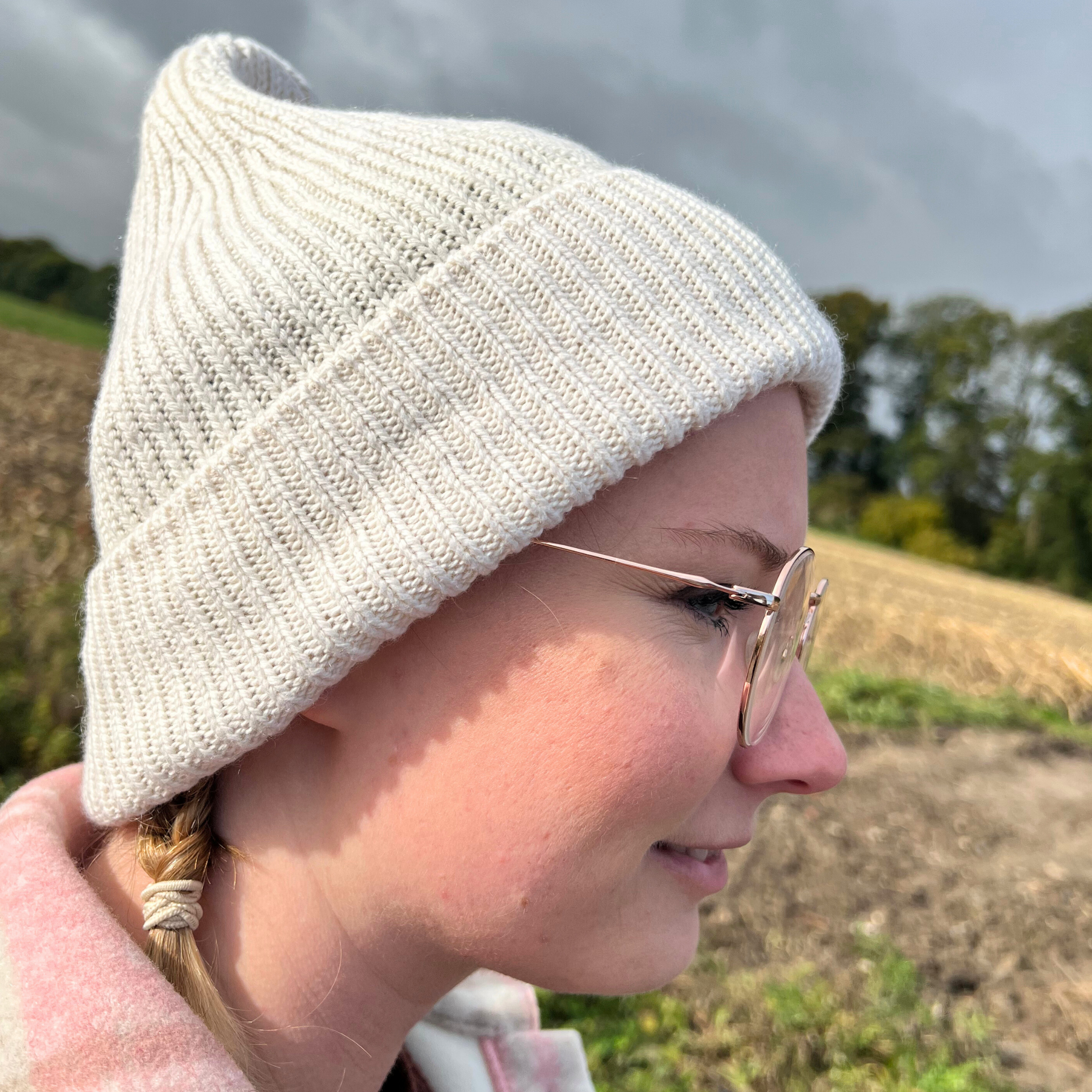Frau mit Brille und weißer Strickmütze im Profil, draußen bei bewölktem Himmel und Feld im Hintergrund.
