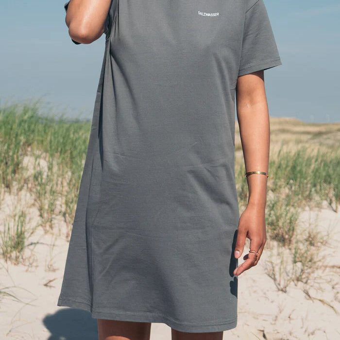 Eine Person in einem locker sitzenden grauen T-Shirt-Kleid steht an einem Sandstrand mit hohem Gras im Hintergrund. Ihr Gesicht ist nicht zu sehen, und sie trägt ein Armband und einen Ring.
