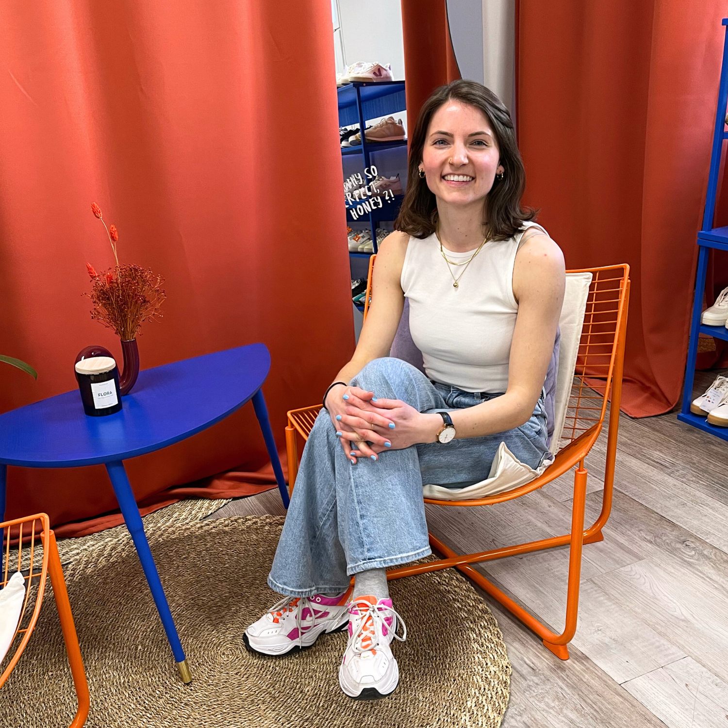 Frau in einem weißen Top und Jeans sitzt lächelnd auf einem Stuhl neben einem Tisch mit einer Vase vor roten Vorhängen.