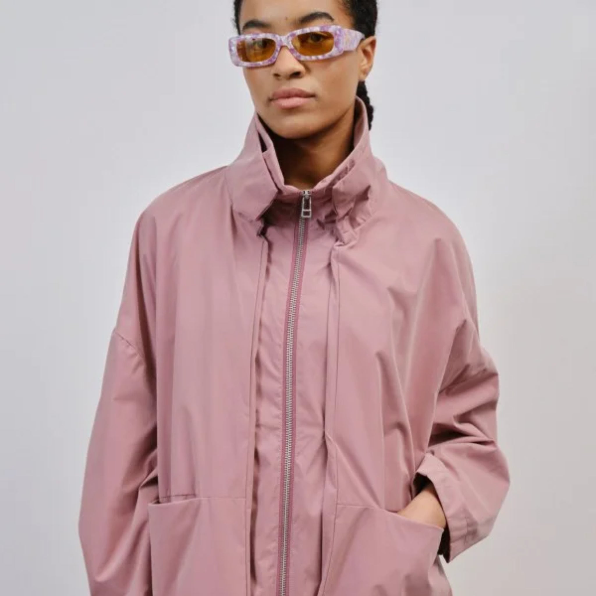 Person in rosa Jacke mit hohem Kragen und gelben Sonnenbrillen, die in die Kamera schaut.