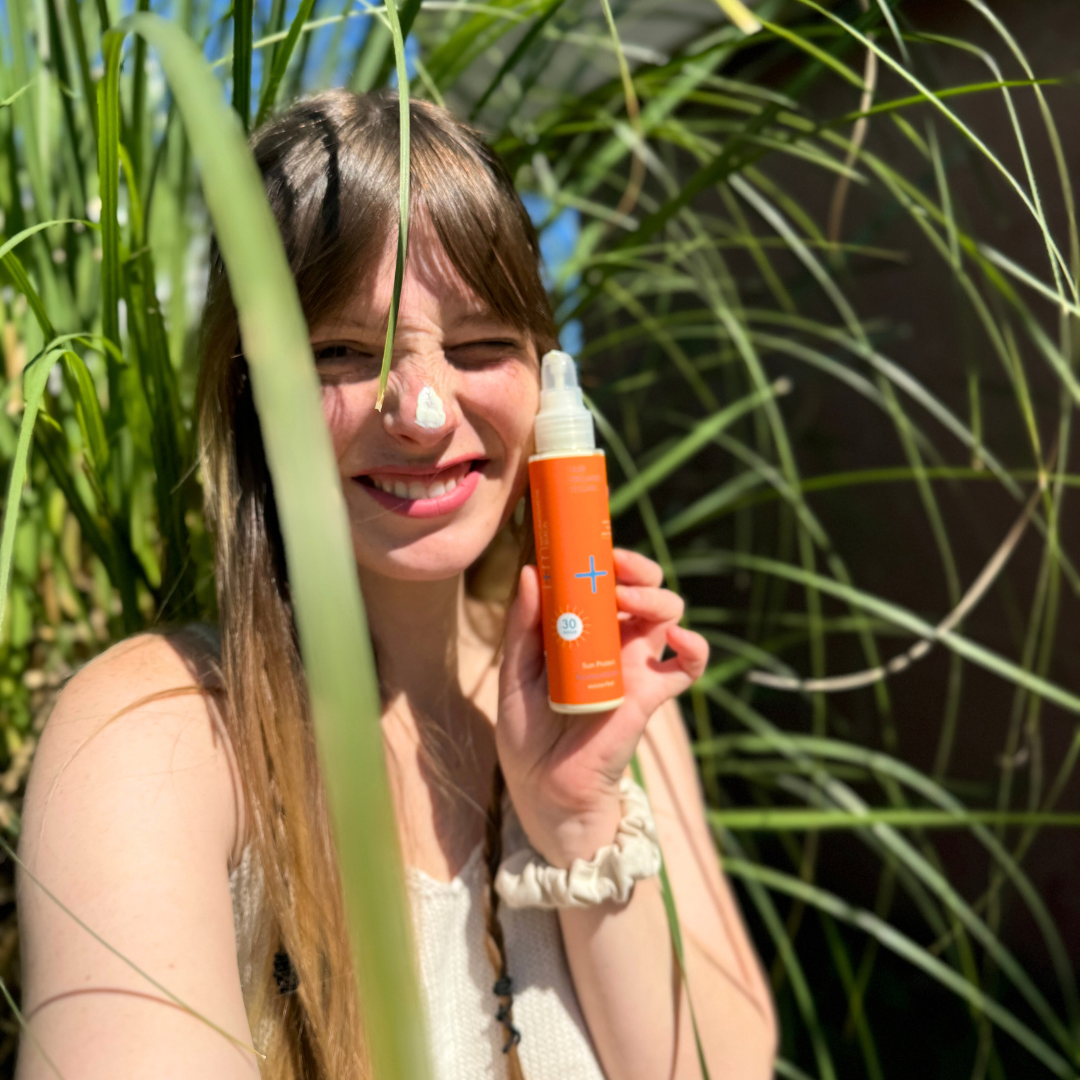Frau mit Sonnencreme auf der Nase hält eine orangefarbene Flasche in der Hand, umgeben von grünen Pflanzen.