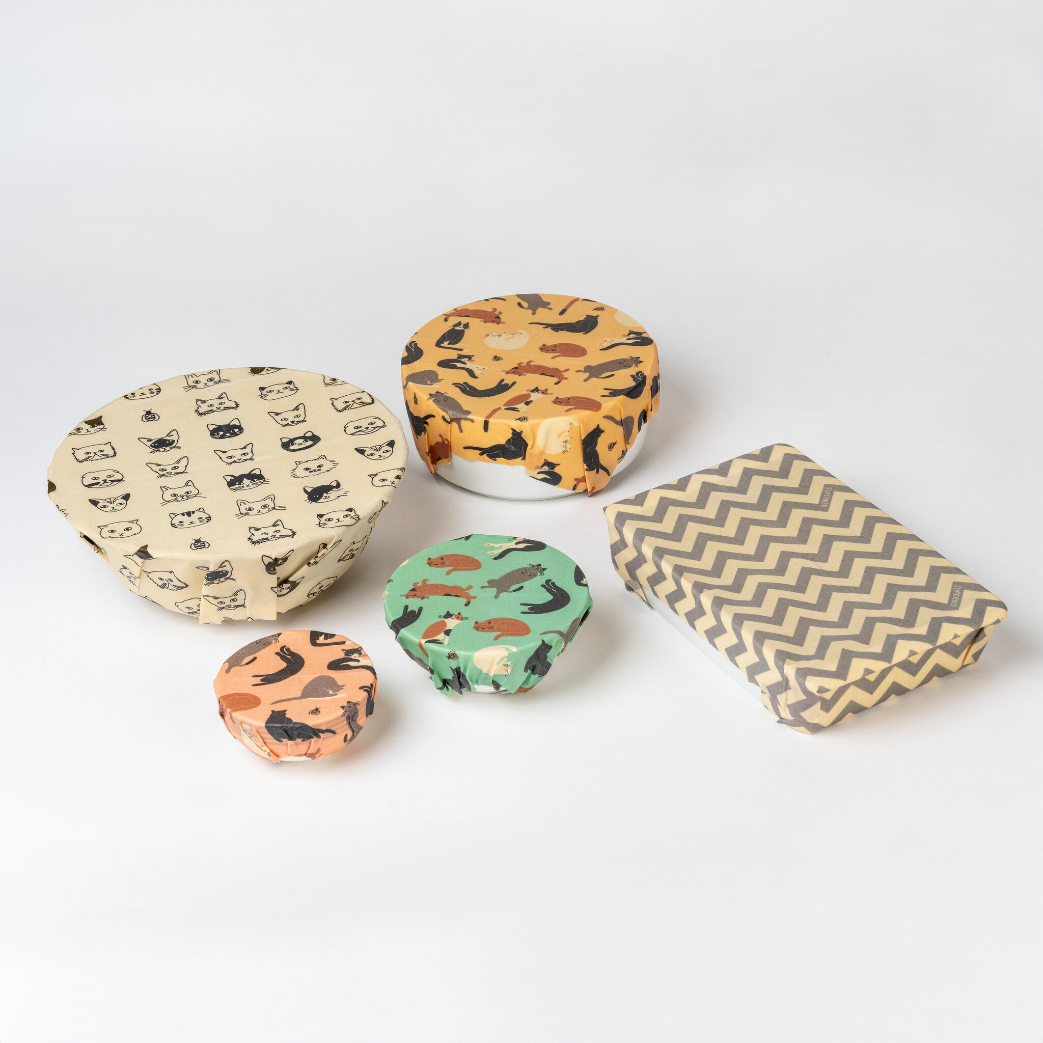 Vier Schalen mit wiederverwendbaren Bienenwachspapieren mit Tiermotiven und ein rechteckiger Gegenstand, der mit einem grau-weißen Bienenwachspapier mit Chevron-Muster umwickelt ist, stehen auf einer hellen Unterlage.