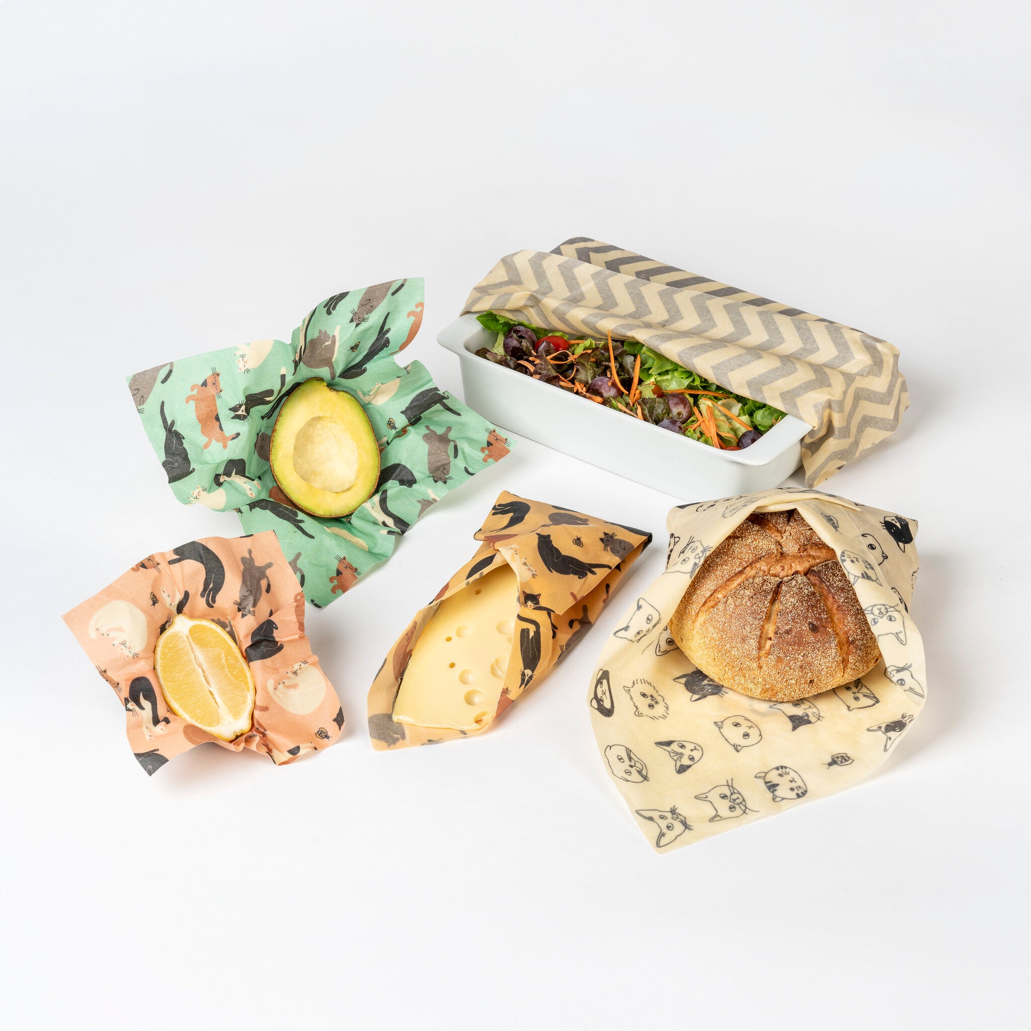 Ein Salat, Brot, Käse, eine halbe Avocado und eine halbe Zitrone sind jeweils in wiederverwendbare gemusterte Bienenwachspackungen auf weißem Hintergrund eingewickelt.