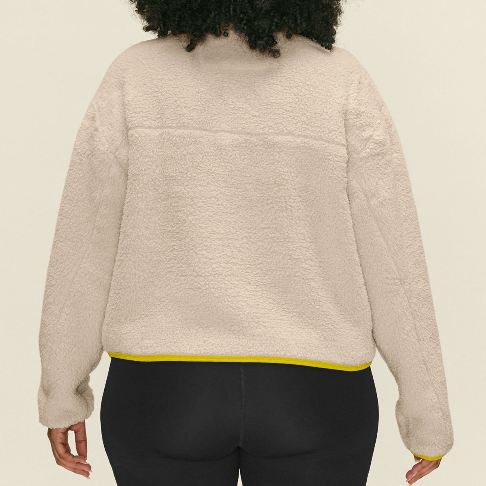 Eine Person mit lockigem Haar, die eine beigefarbene Fleecejacke mit gelbem Saum und schwarze Leggings trägt, von hinten vor einem schlichten hellen Hintergrund dargestellt.