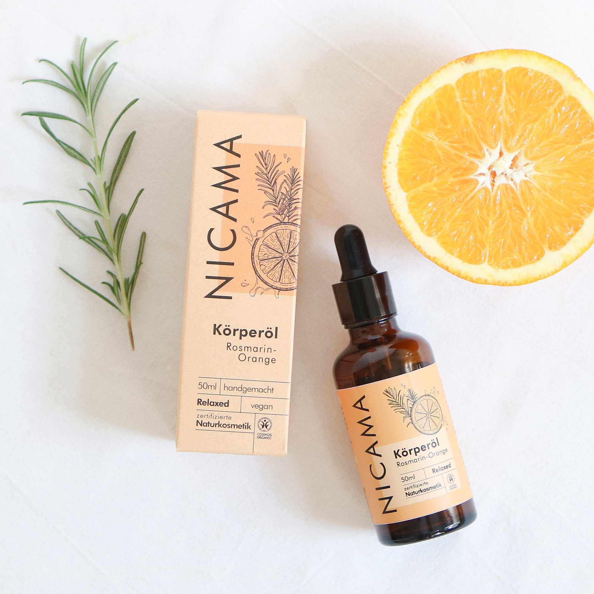 Flasche und Verpackung von NICAMA Körperöl mit Rosmarin-Zweig und halber Orange auf weißem Hintergrund.