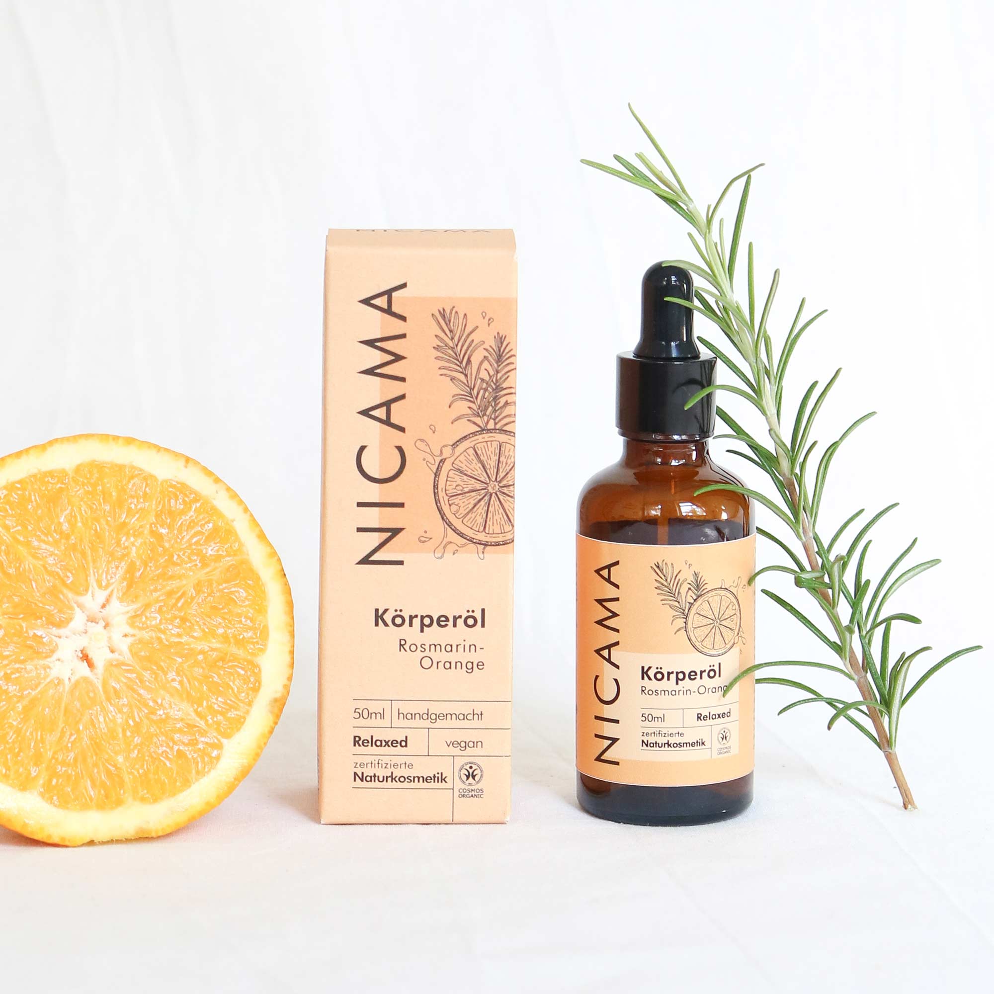 Flasche und Verpackung von Nicama Körperöl Rosmarin-Orange mit halber Orange und Rosmarinzweig auf weißem Hintergrund.