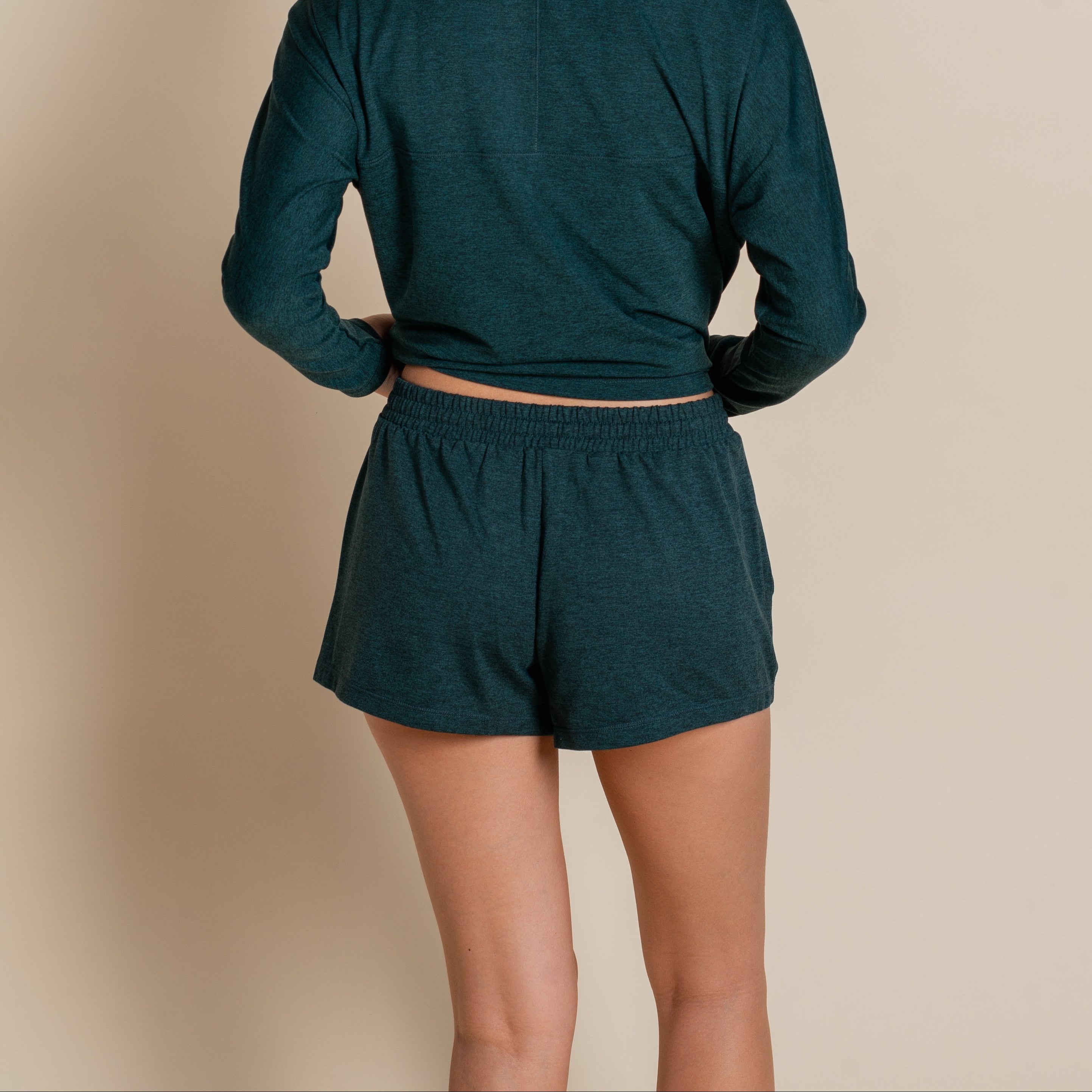 Eine Frau steht von der Kamera abgewandt in einem dunkelgrünen, langärmeligen Oberteil und einer kurzen Hose. Ihre rechte Hand ruht auf ihrer Hüfte, und sie hat schulterlanges braunes Haar.