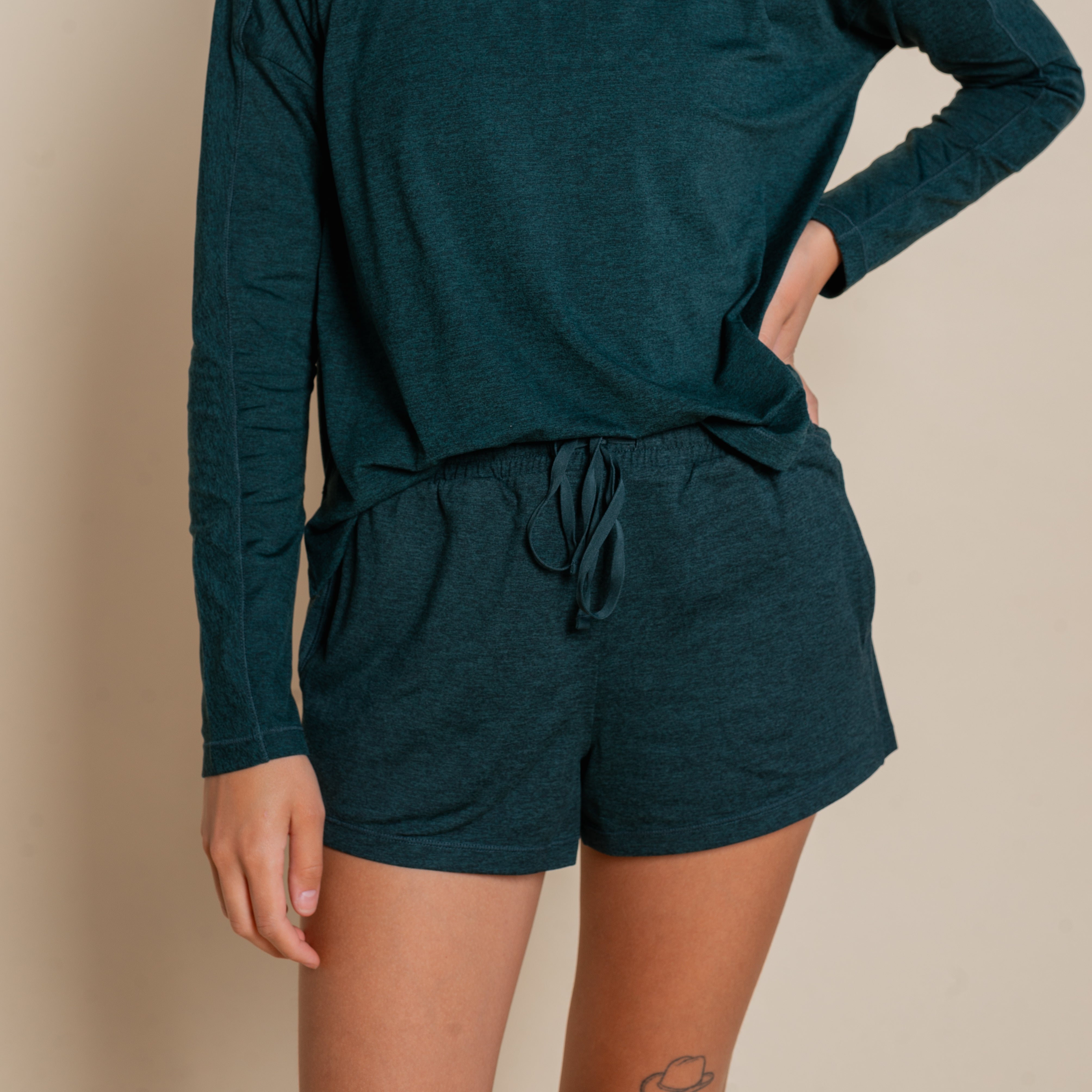 Eine Frau in einem dunkelgrünen, langärmeligen Hemd und passenden Shorts mit Kordelzug steht vor einem beigen Hintergrund und hat eine Hand an der Hüfte.