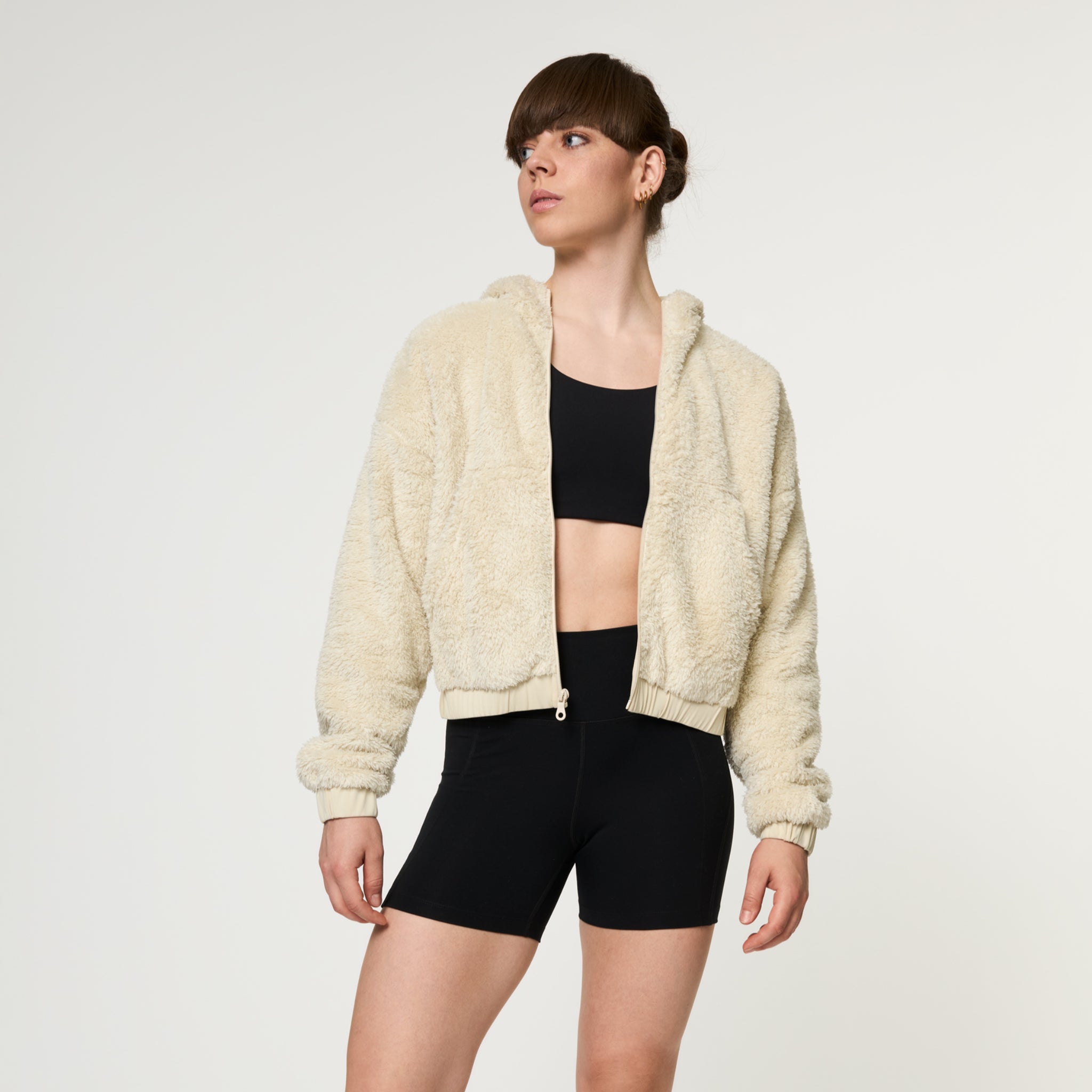 Eine Frau, die eine cremefarbene, wattierte Jacke mit Reißverschluss über einem schwarzen Sport-BH und schwarzen Radlerhosen trägt, steht vor einem schlichten weißen Hintergrund und blickt zur Seite.