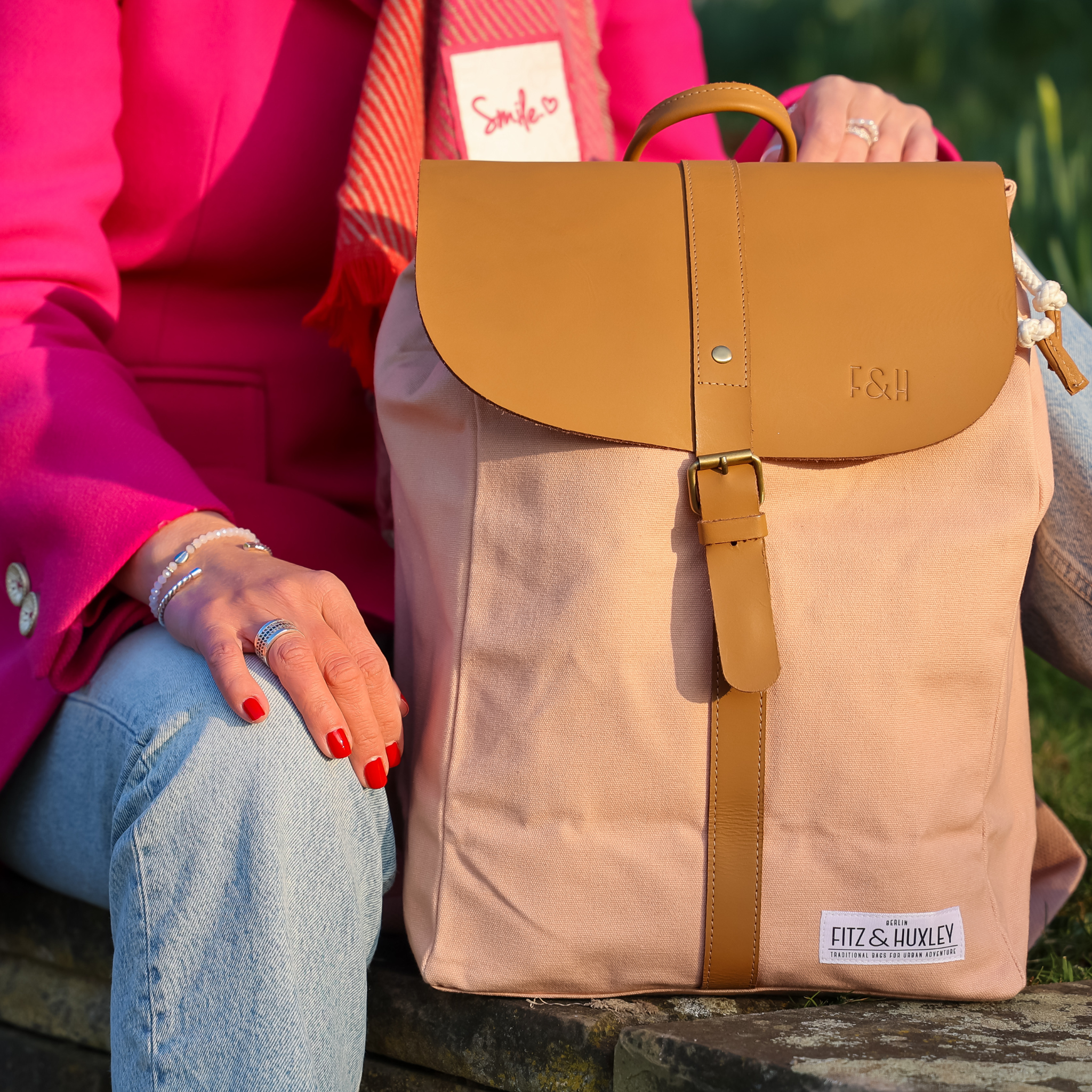 Person mit pinkem Mantel sitzt neben einem beigen Rucksack mit braunem Lederverschluss.