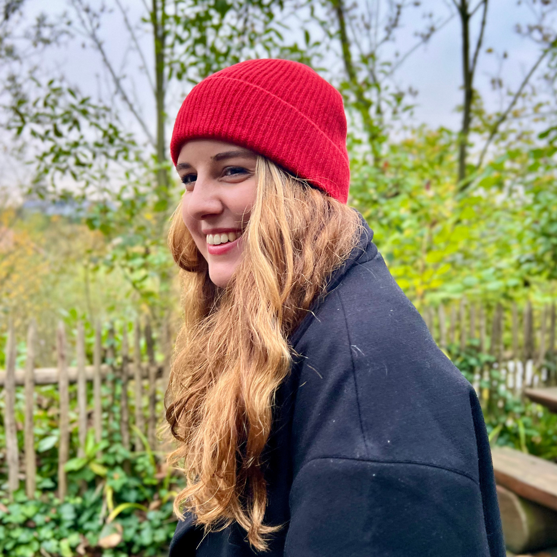 Frau mit roter Mütze und langen Haaren lächelt im Freien, umgeben von Pflanzen und einem Holzzaun.