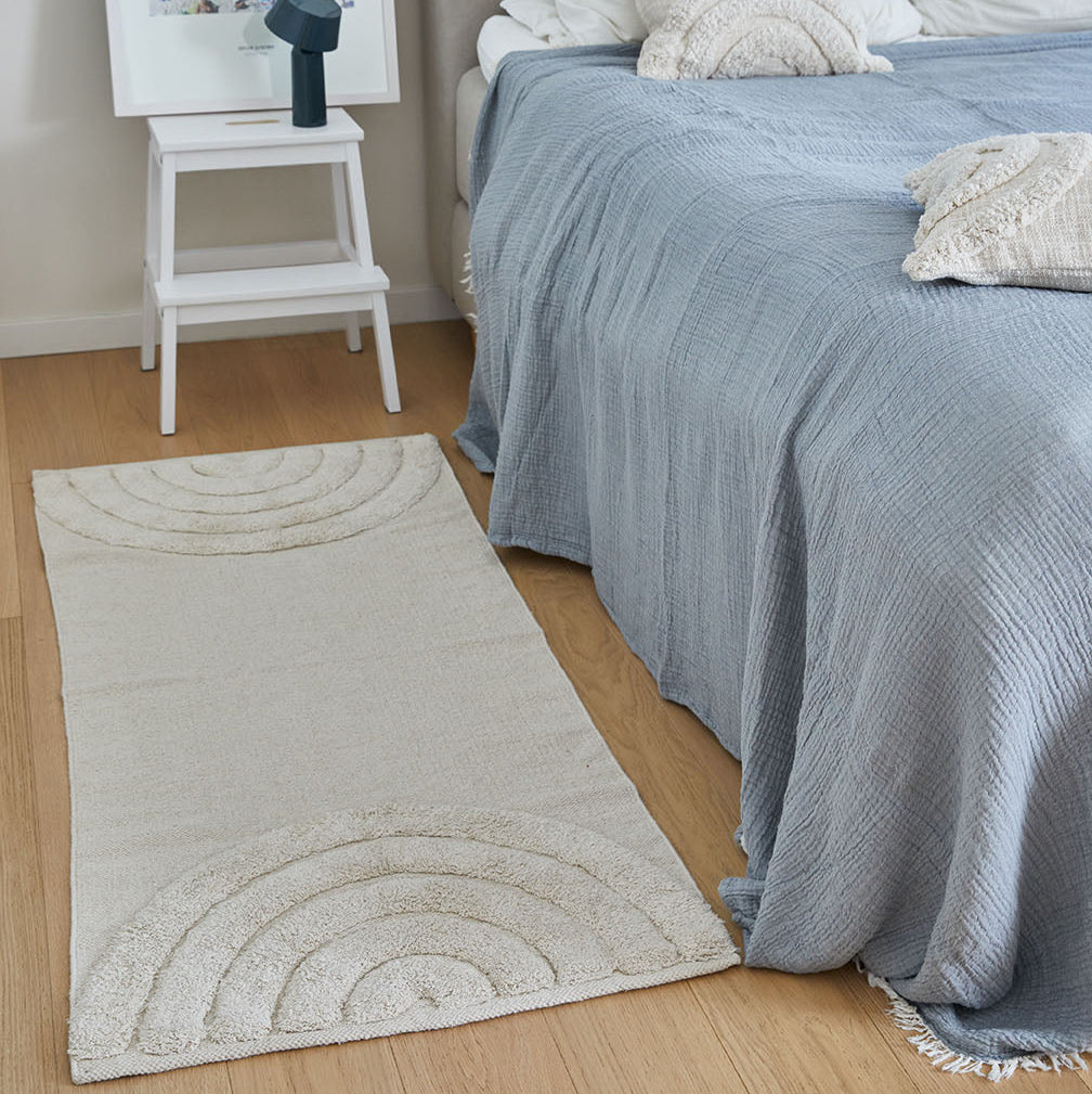 Beige Teppich mit halbkreisförmigem Muster neben einem Bett mit blauer Tagesdecke und einem weißen Nachttisch.