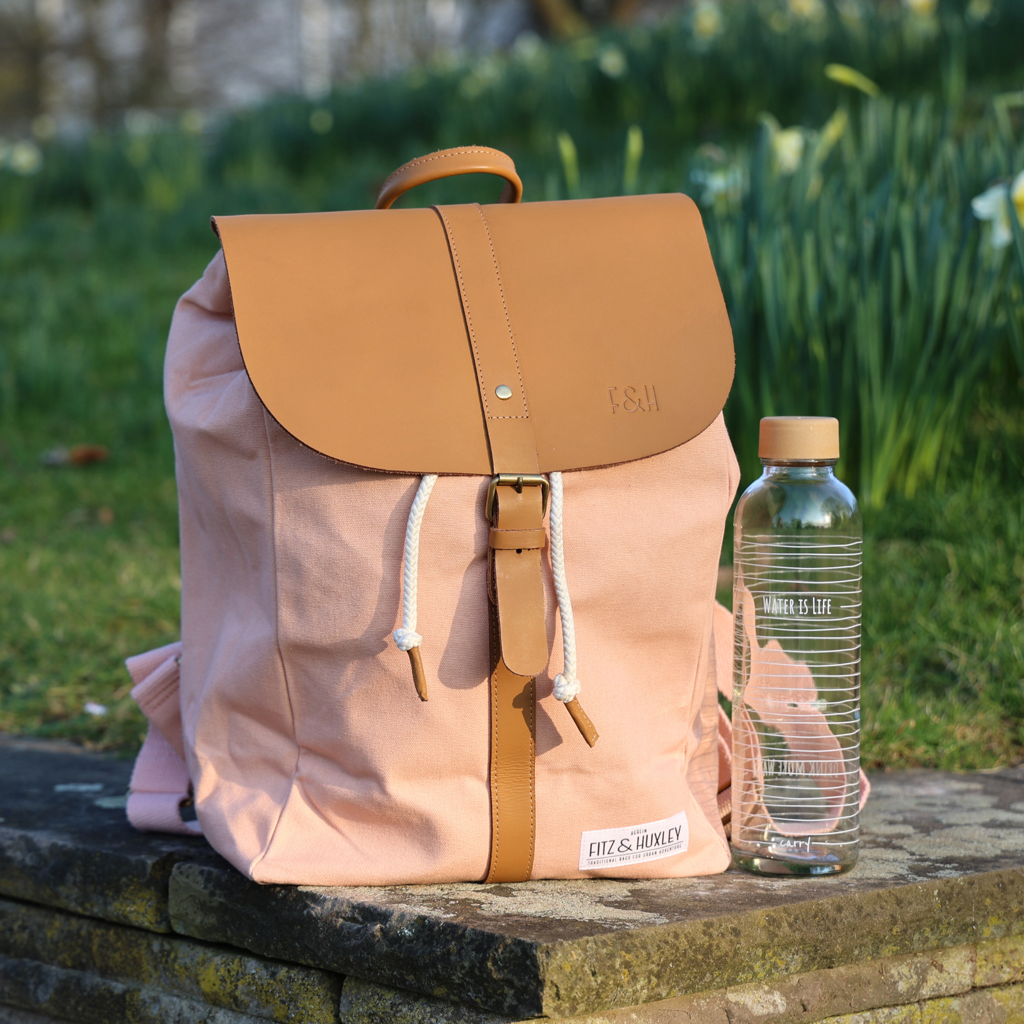 Ein rosa Rucksack mit braunen Lederakzenten und eine Wasserflasche stehen auf einer Steinmauer im Freien.