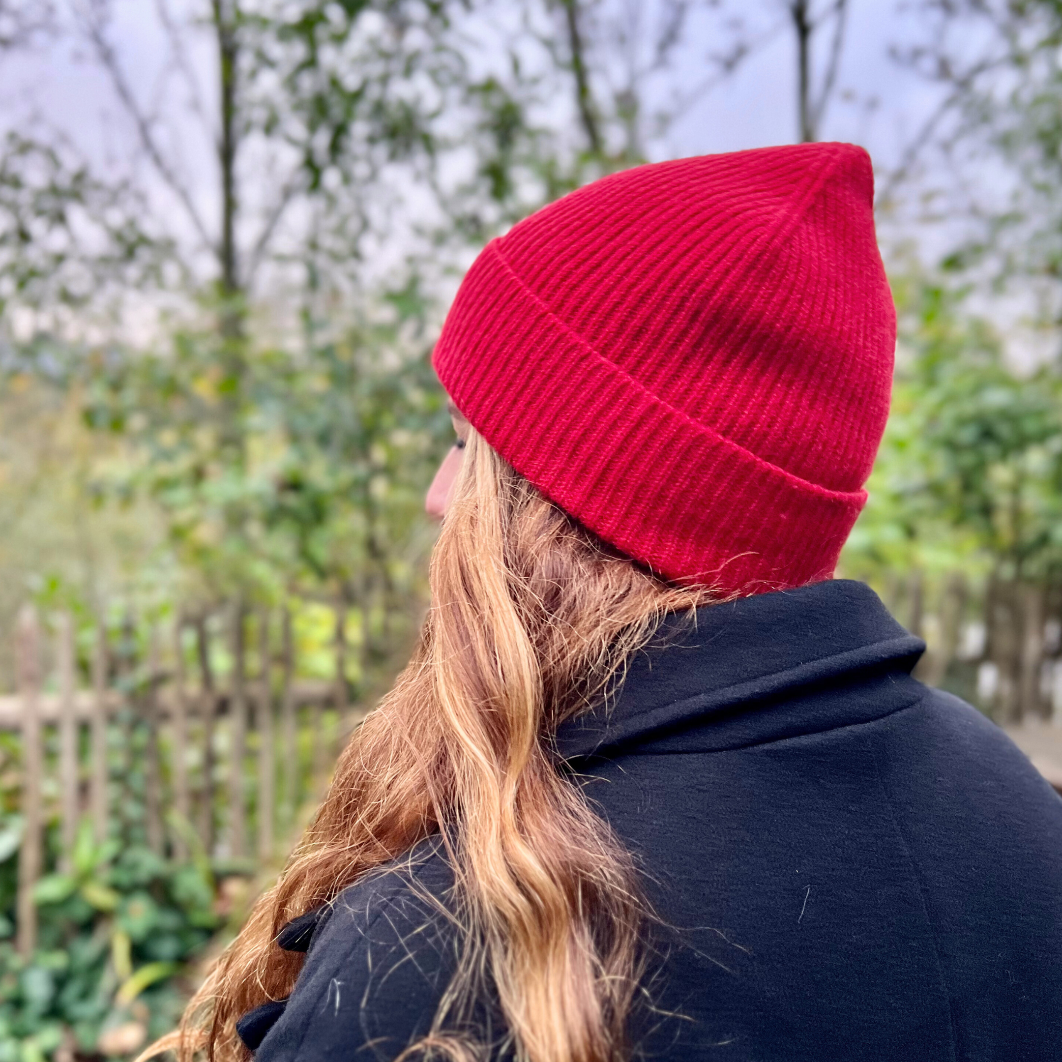 Frau mit roter Mütze und langen, blonden Haaren blickt im Freien auf eine verschwommene, grüne Landschaft.